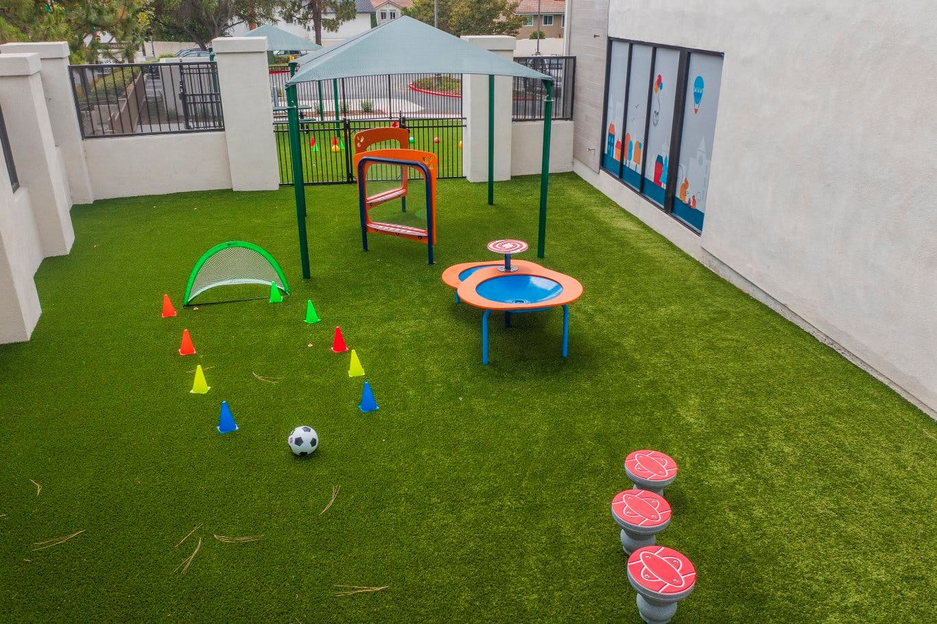 An outdoor play area with artificial turf, a small soccer goal, cones, and play structures