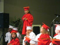 Child in red graduation gown standing, other children in gowns seated