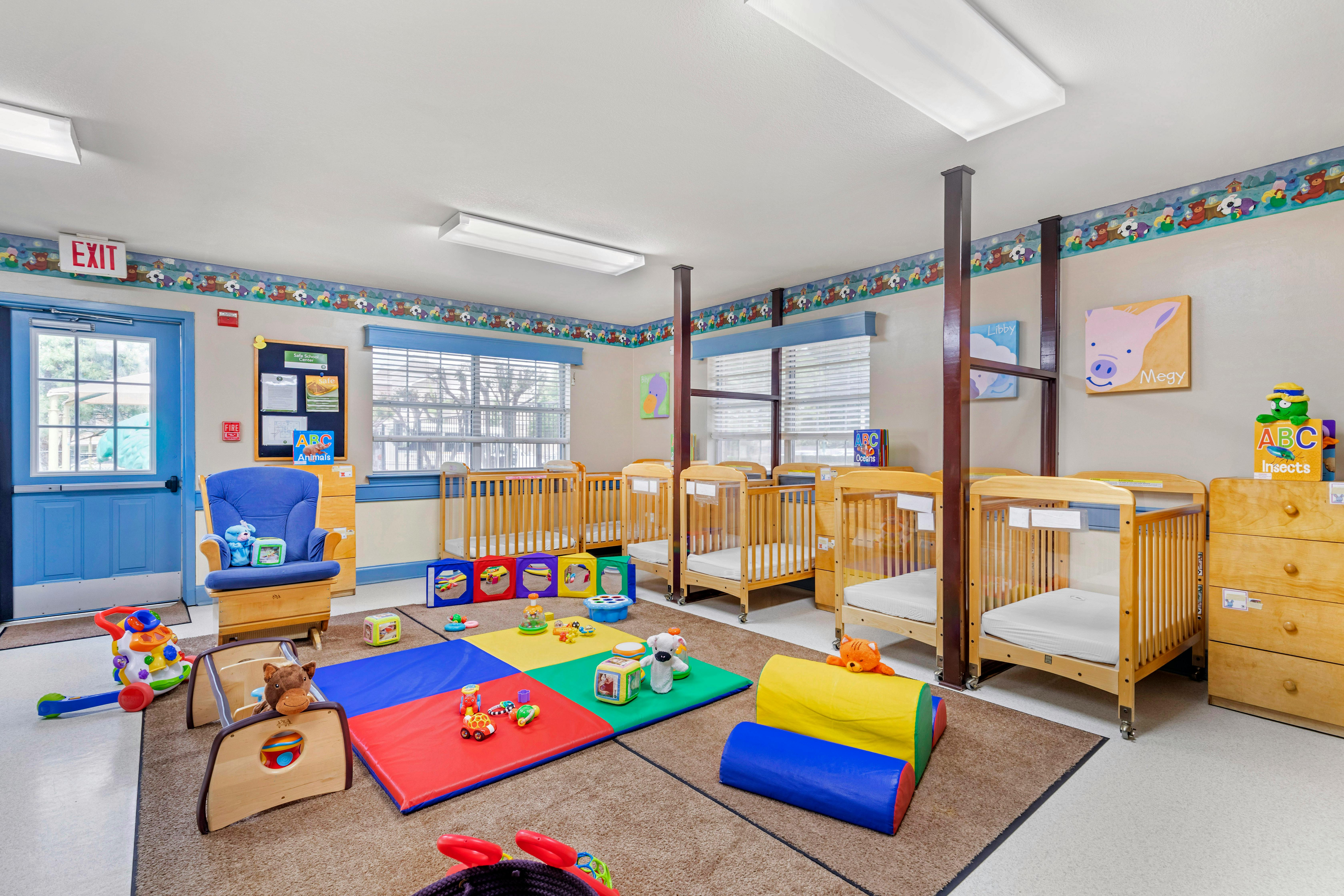 A playroom with six cribs, a blue rocking chair, and colorful playmats and toys