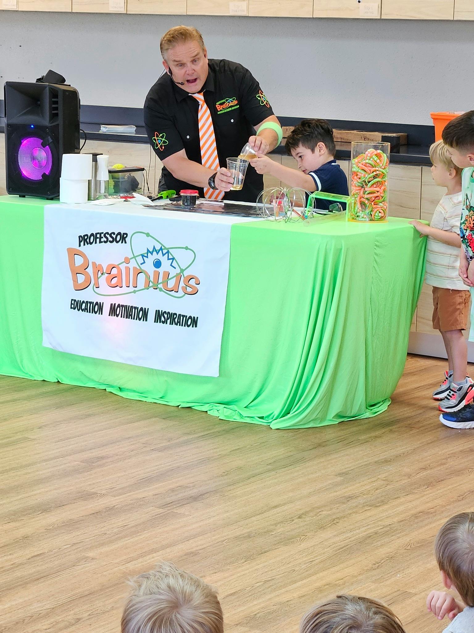 A person pouring liquid into a child's beaker on a table with a "Professor Brainius" sign