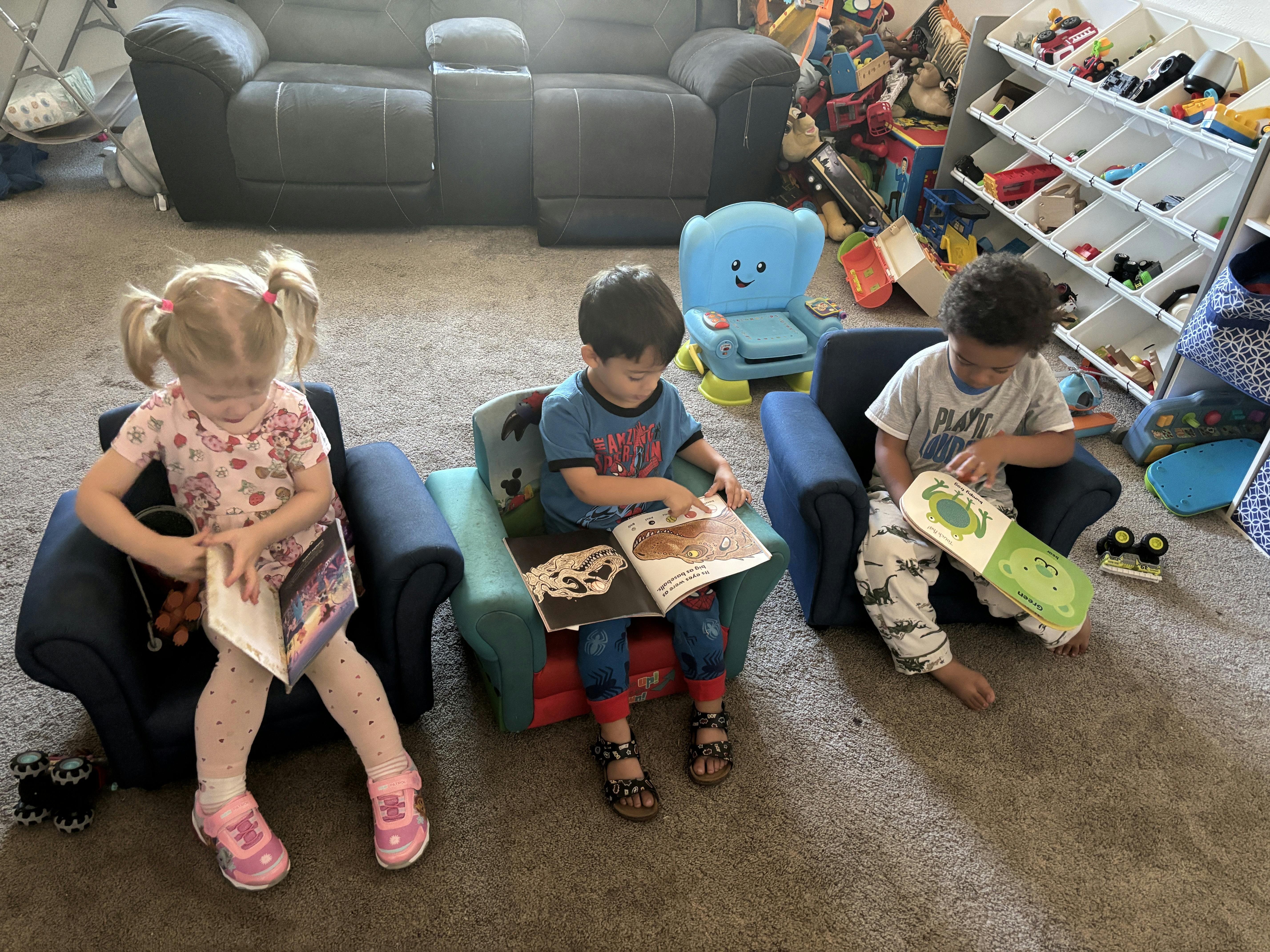 Three children sitting in small chairs reading books in a playroom
