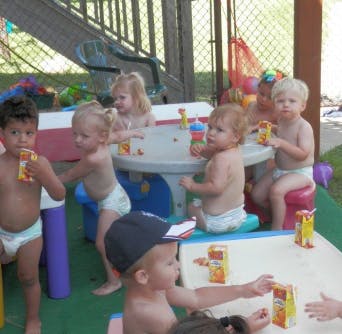 Multiple children in diapers playing with juice boxes at tables outdoors