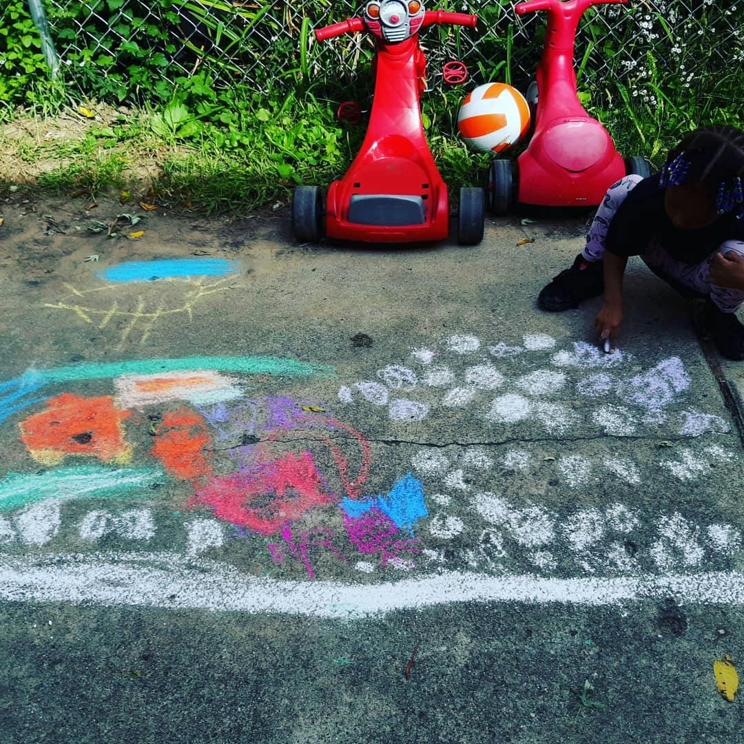 Child drawing with chalk on a sidewalk next to two red ride-on toys