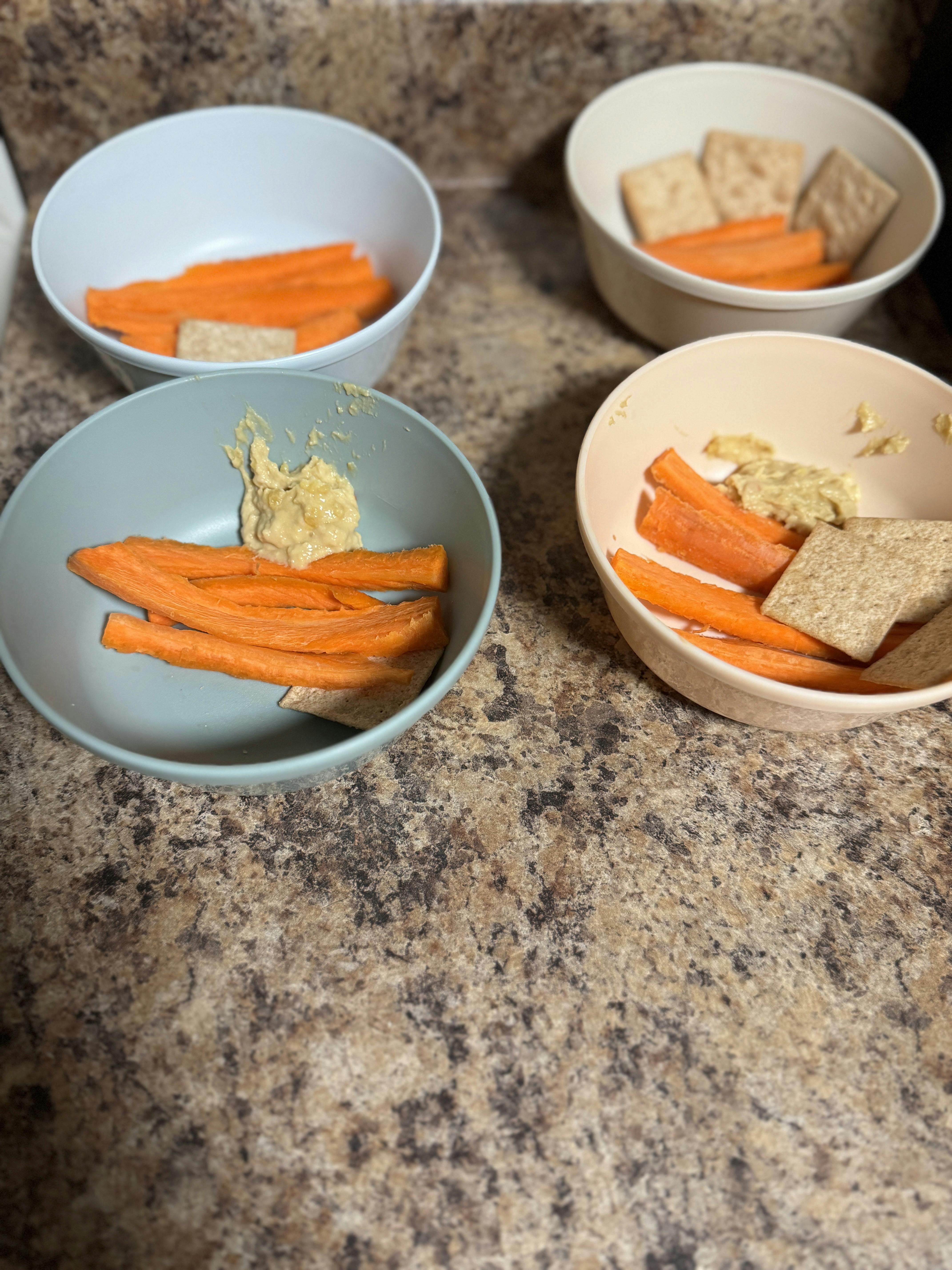 Four bowls containing carrot sticks, crackers, and hummus