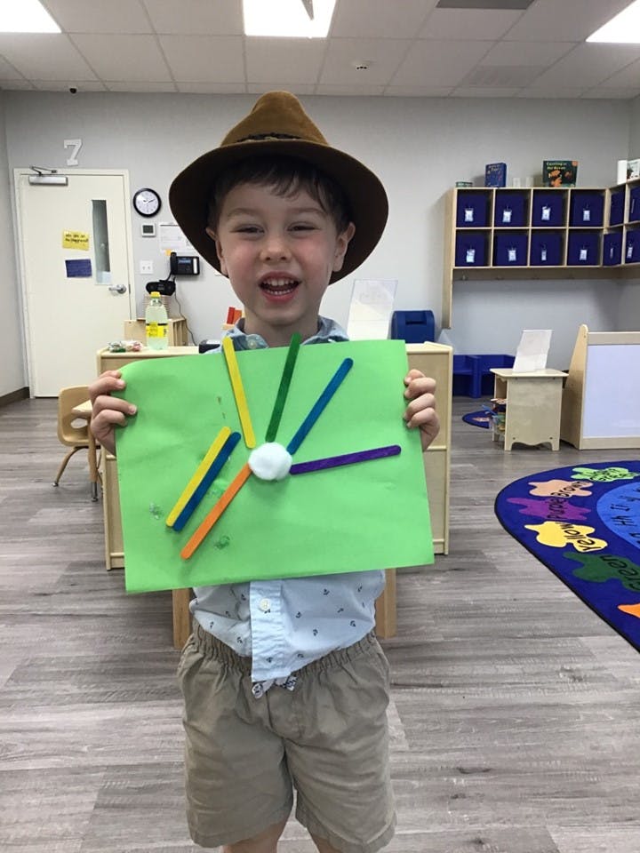 Child in a hat holding up a green paper craft with colorful sticks