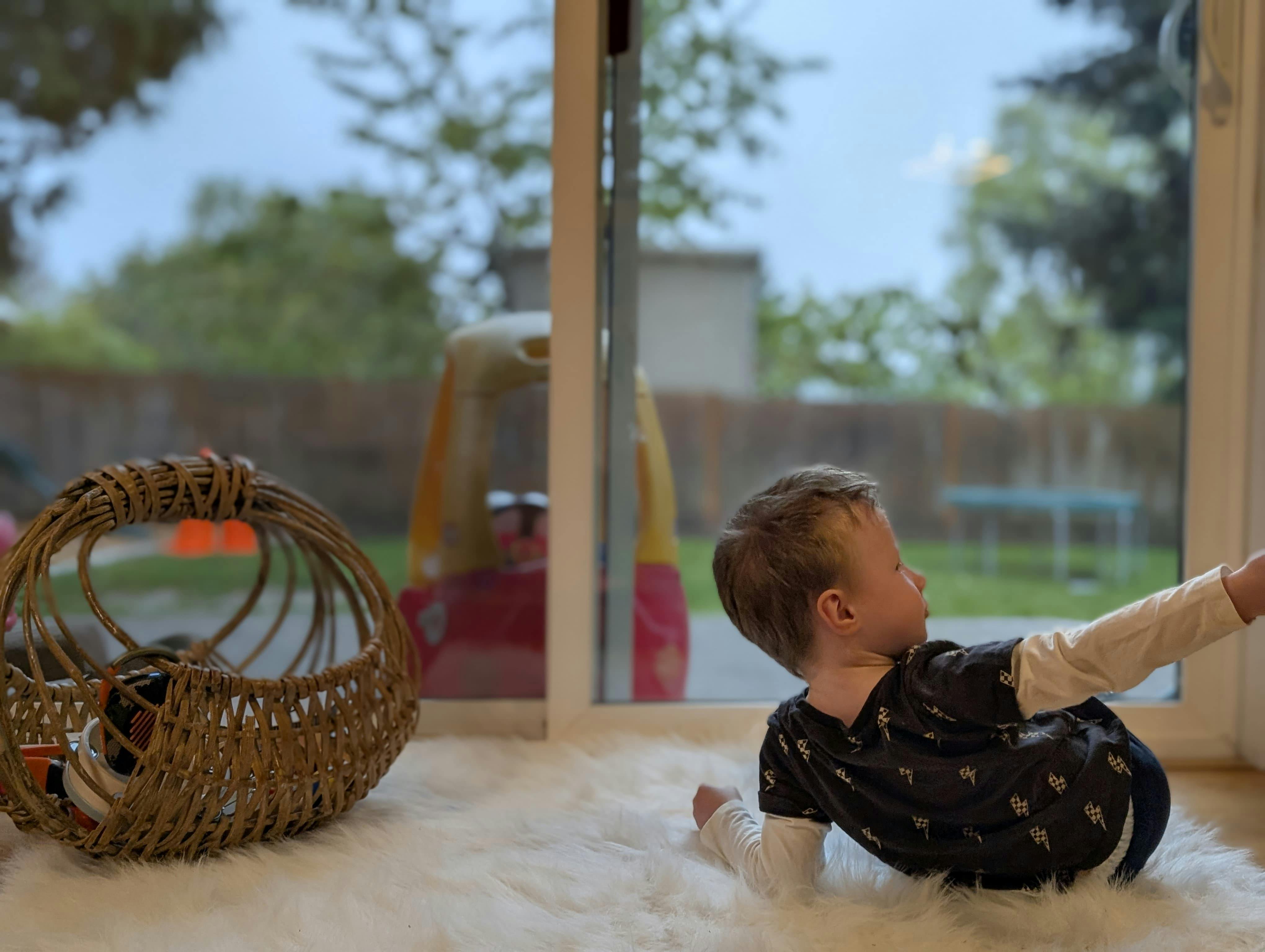 Child kneeling on a white rug next to a woven basket, looking out a glass door
