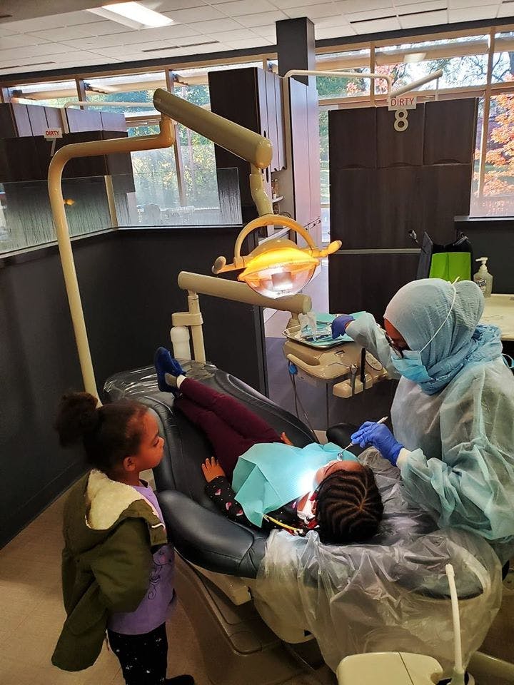 Person in medical attire examining a child in a dental chair, another child watching