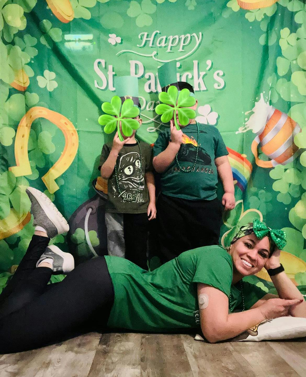 Person lying on floor with two children holding shamrocks in front of faces
