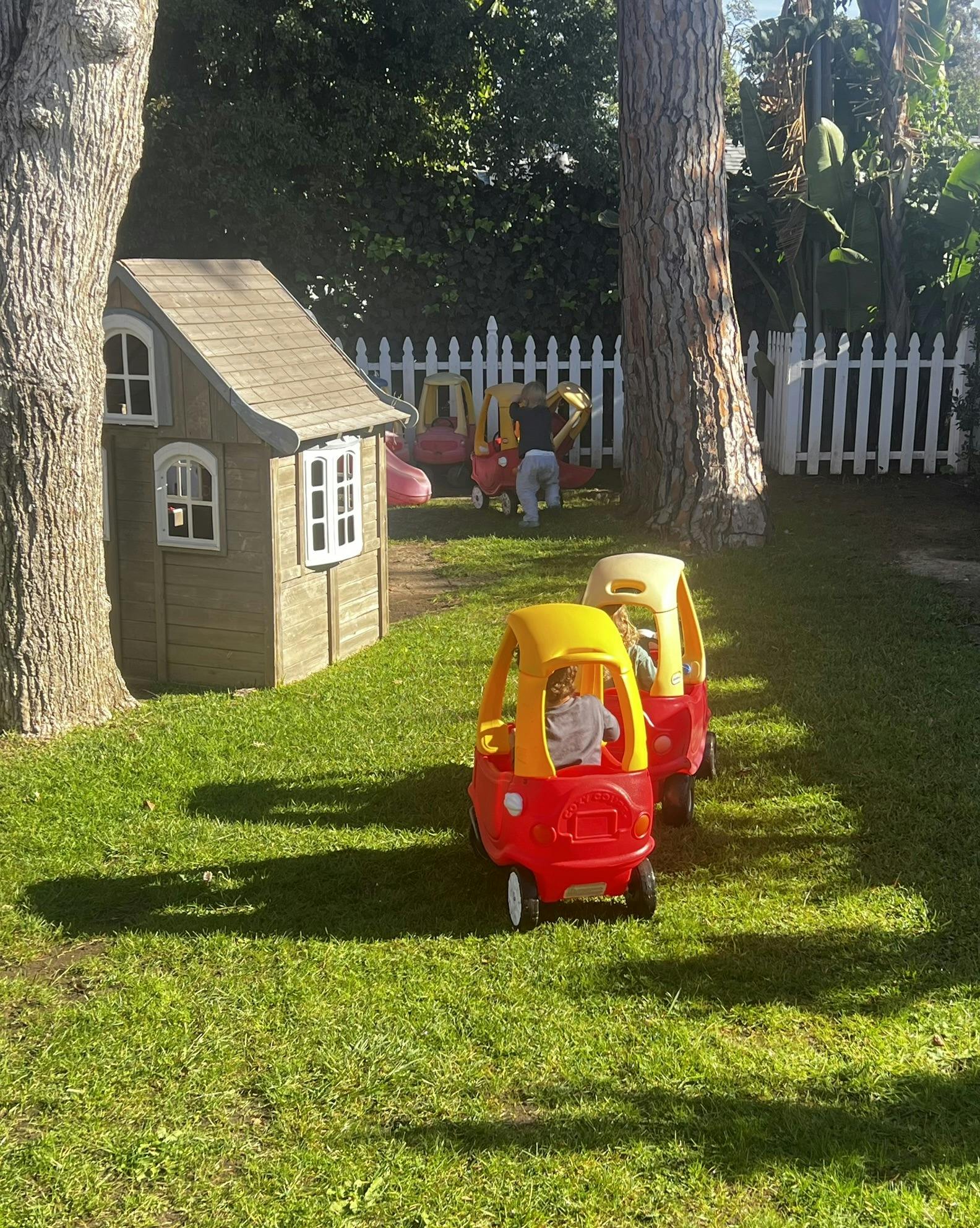 Children playing with toy cars in a grassy backyard with a playhouse