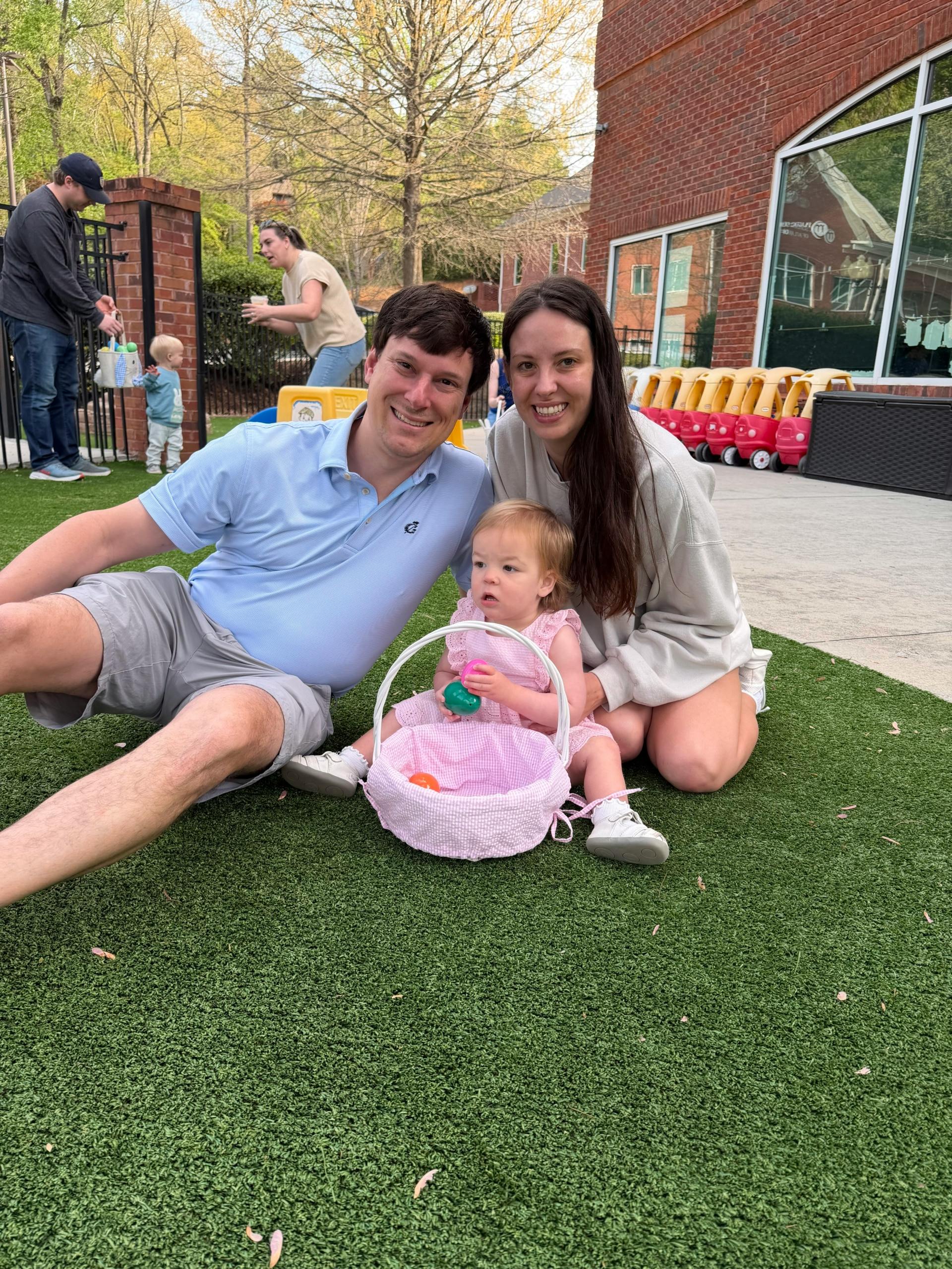 Two adults sitting on grass with a child holding a basket of plastic eggs