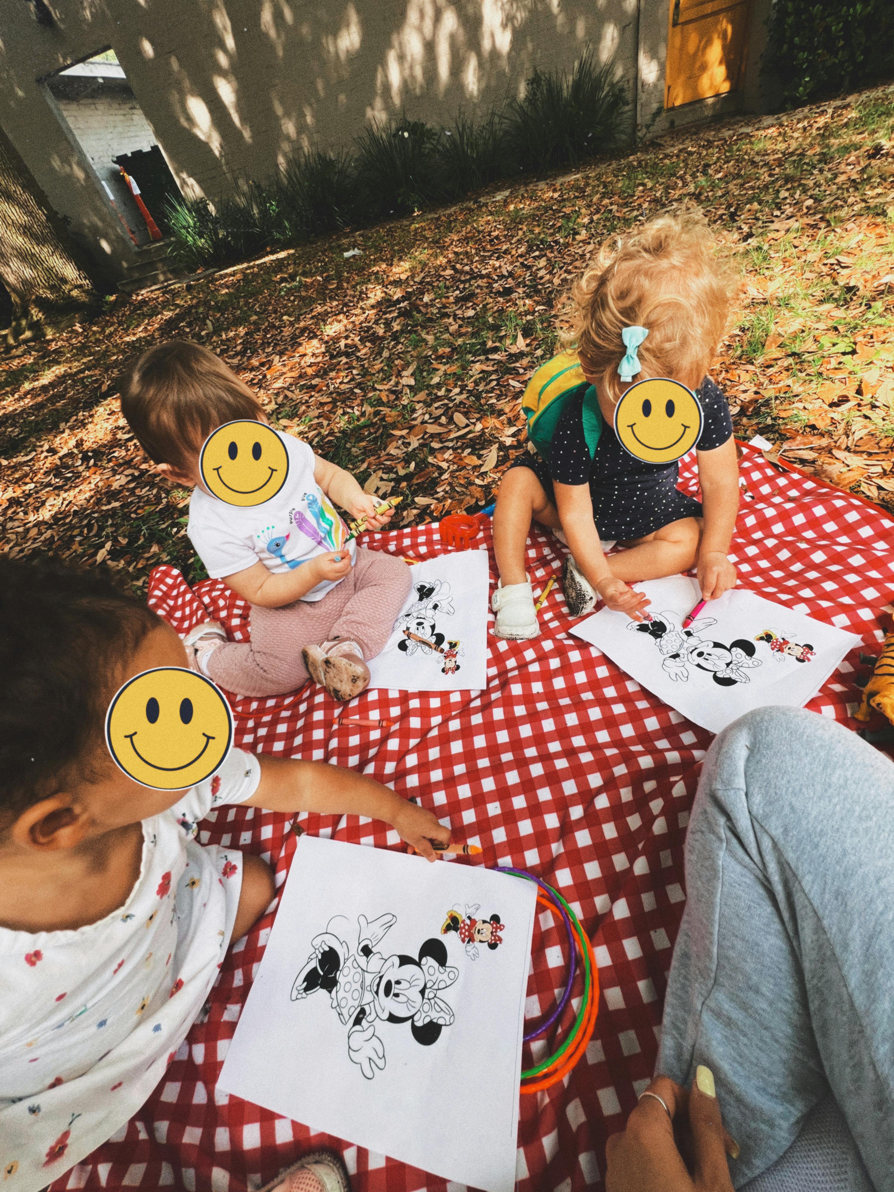 Three children sitting on a red and white checkered blanket, coloring