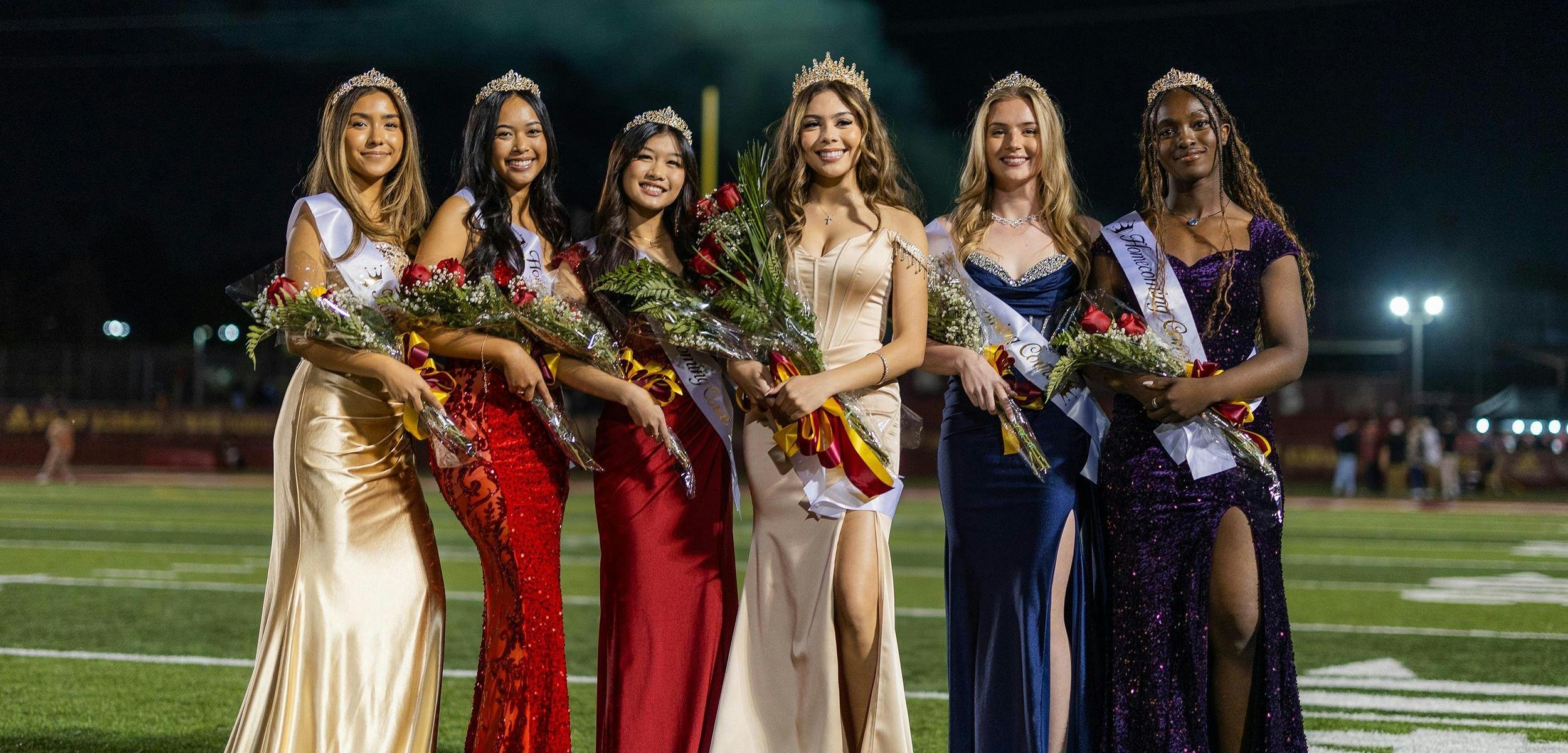 Six teens wearing crowns, sashes, and formal dresses, holding bouquets on a football field