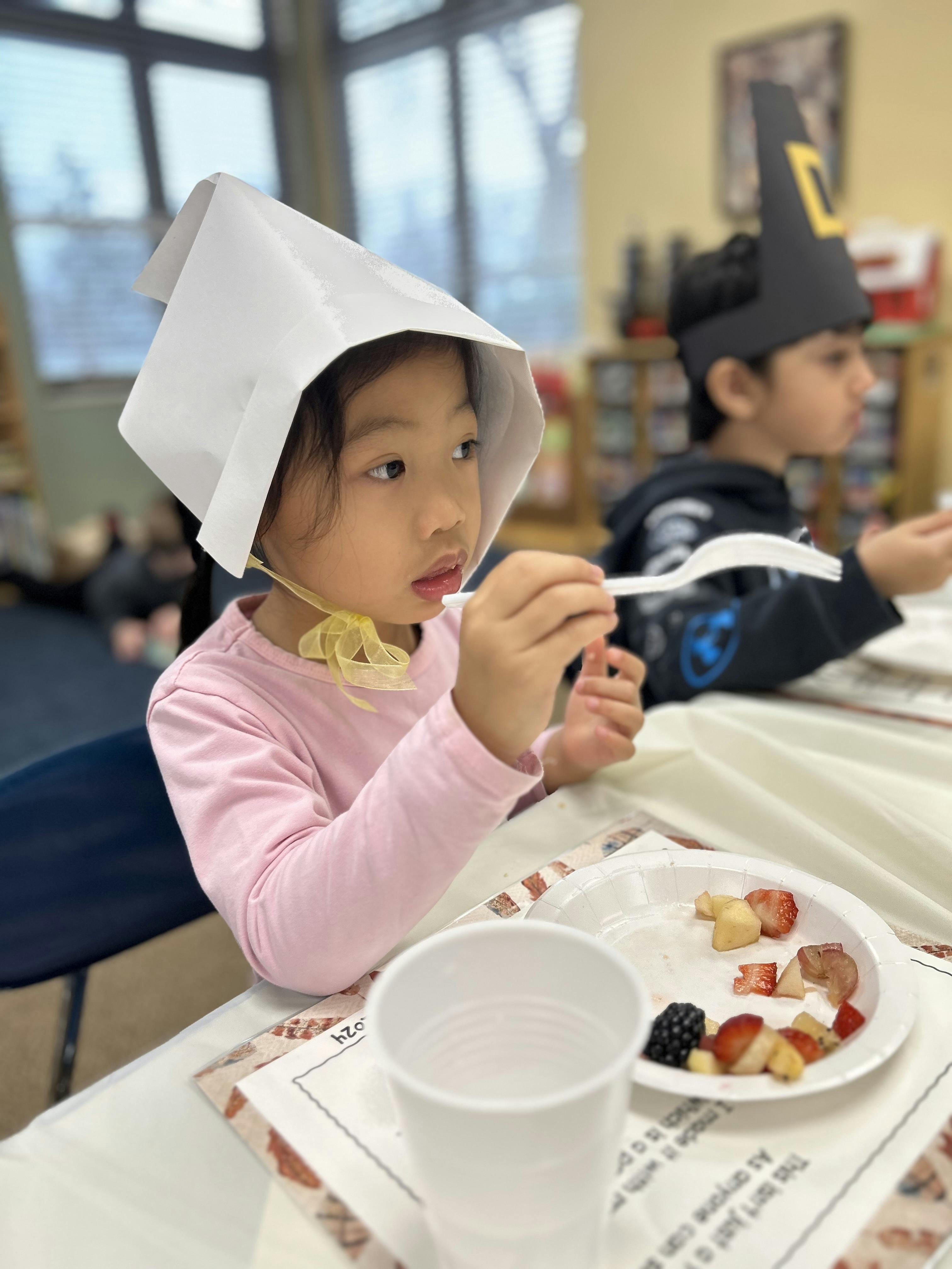 Child in a white paper hat eating fruit from a plate with a plastic fork