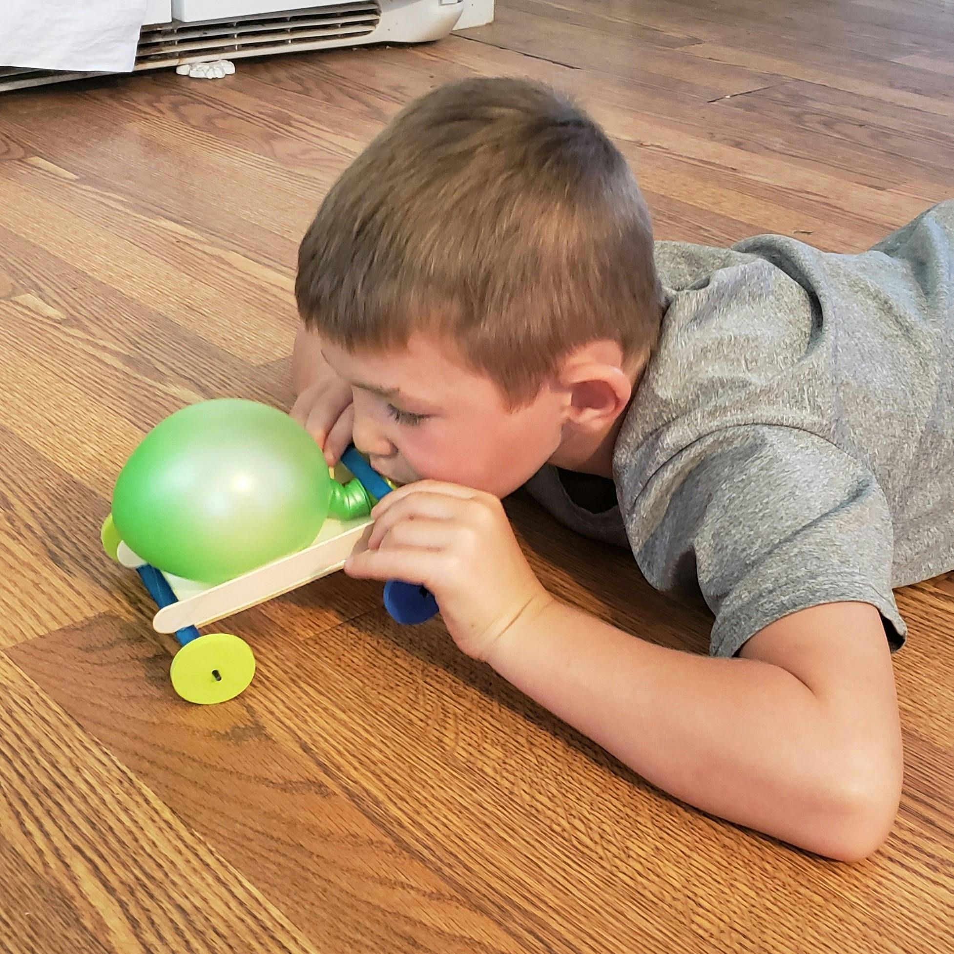 Child blowing up a balloon attached to a small wooden car on the floor
