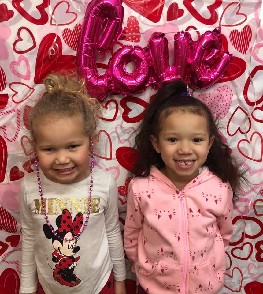 Two children smiling in front of a heart-patterned background with "LOVE" balloons