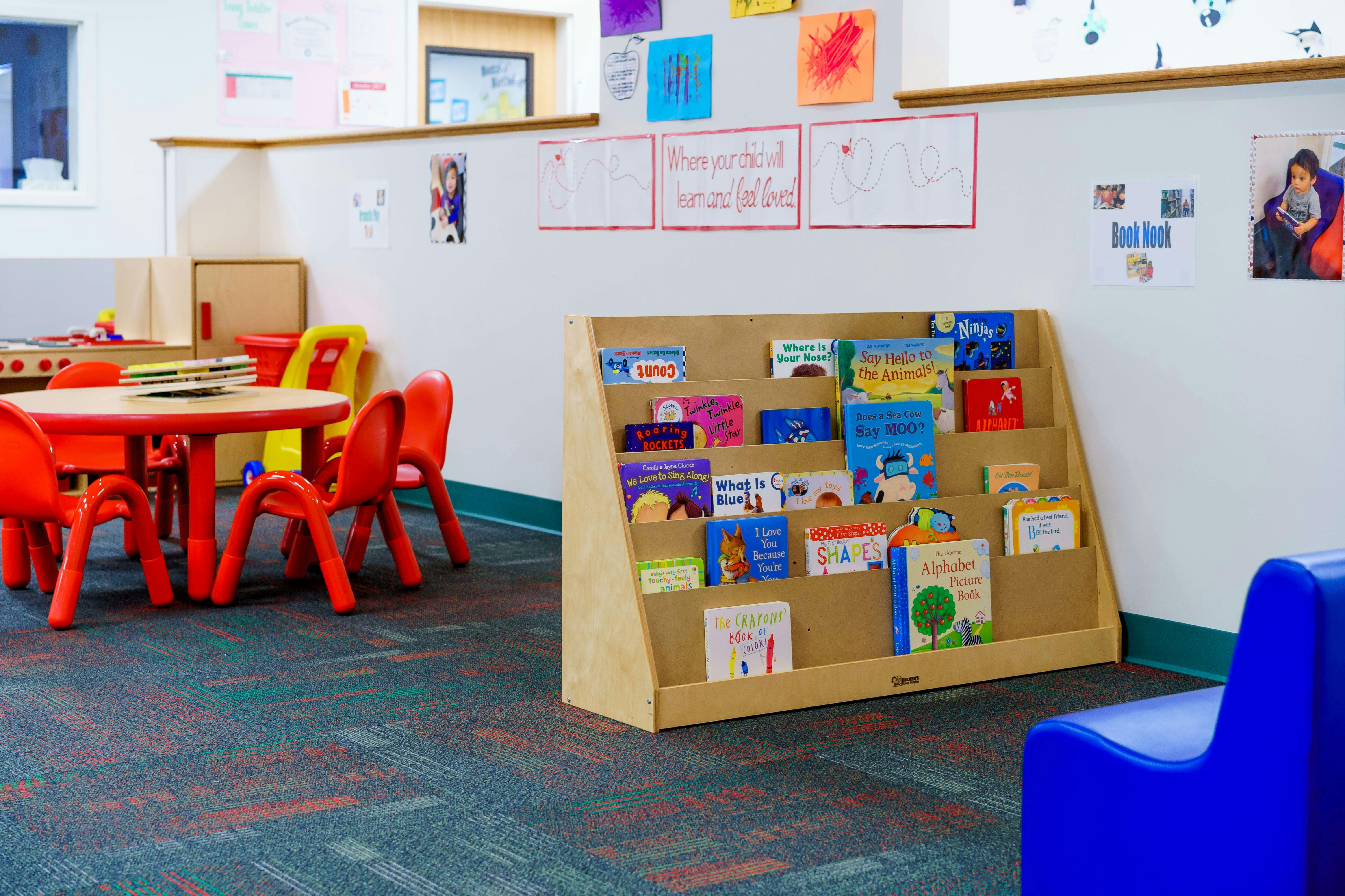 Children's books on a wooden shelf in a classroom with a red table and chairs