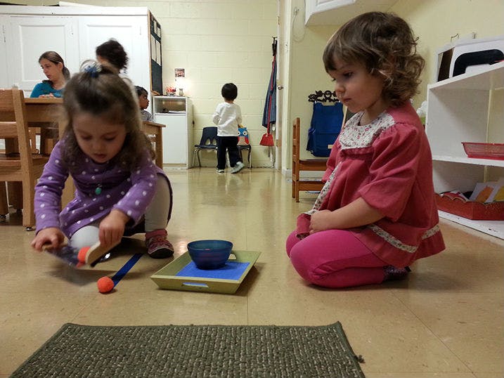 Child sweeping a mat with a brush and dustpan, another child sitting nearby