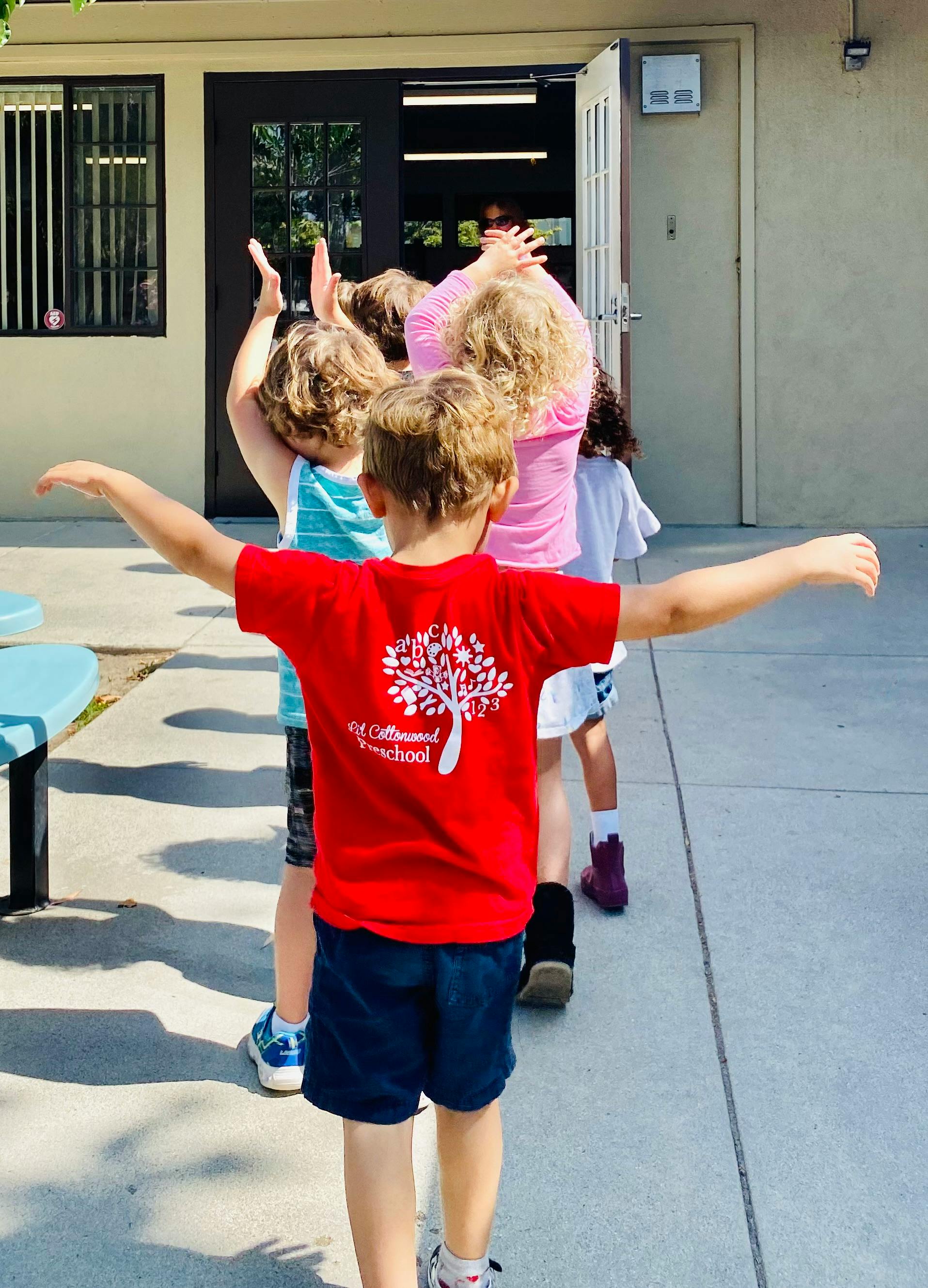 Children standing in a line with arms raised, some raised, outside a building