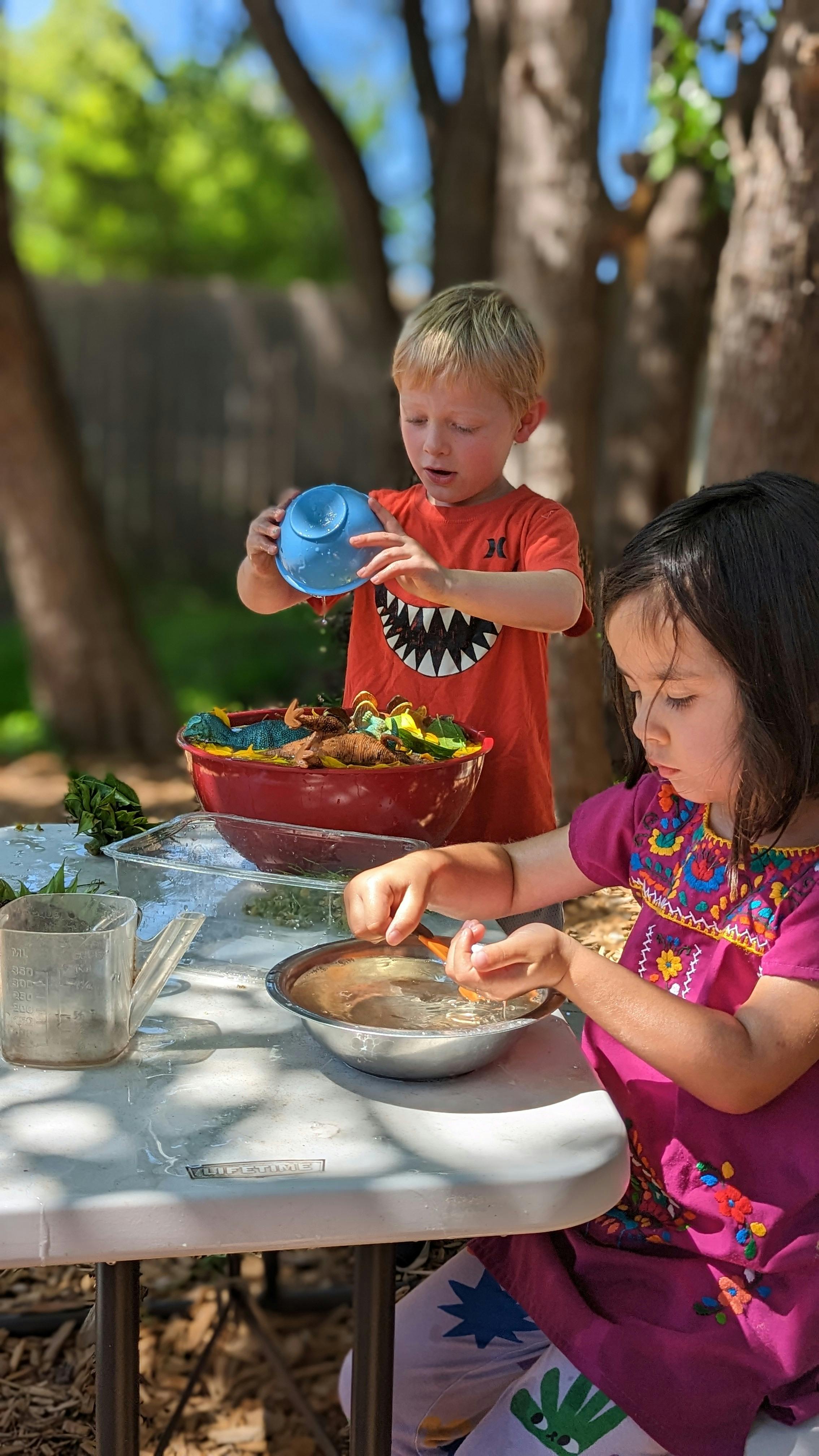 Child pouring water from a blue cup into a red bowl, another child stirring a bowl