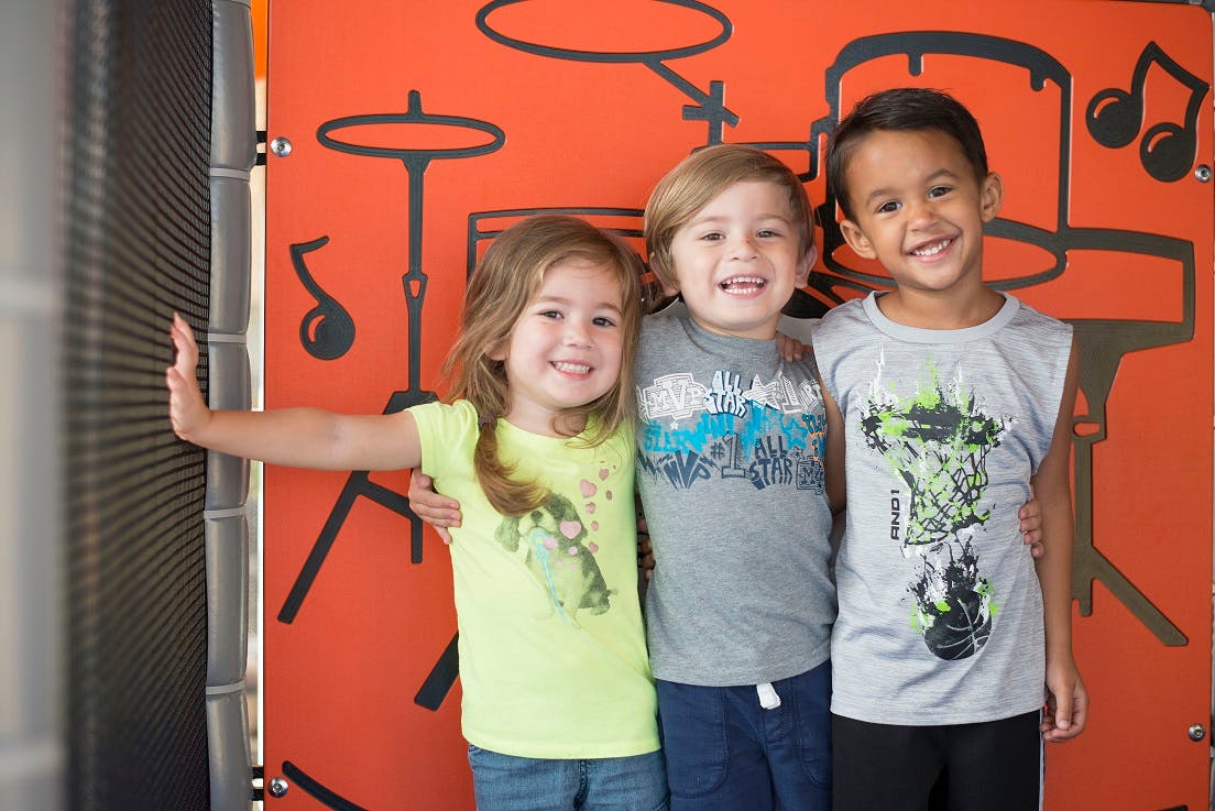 Three children standing together in front of a wall with musical instrument drawings