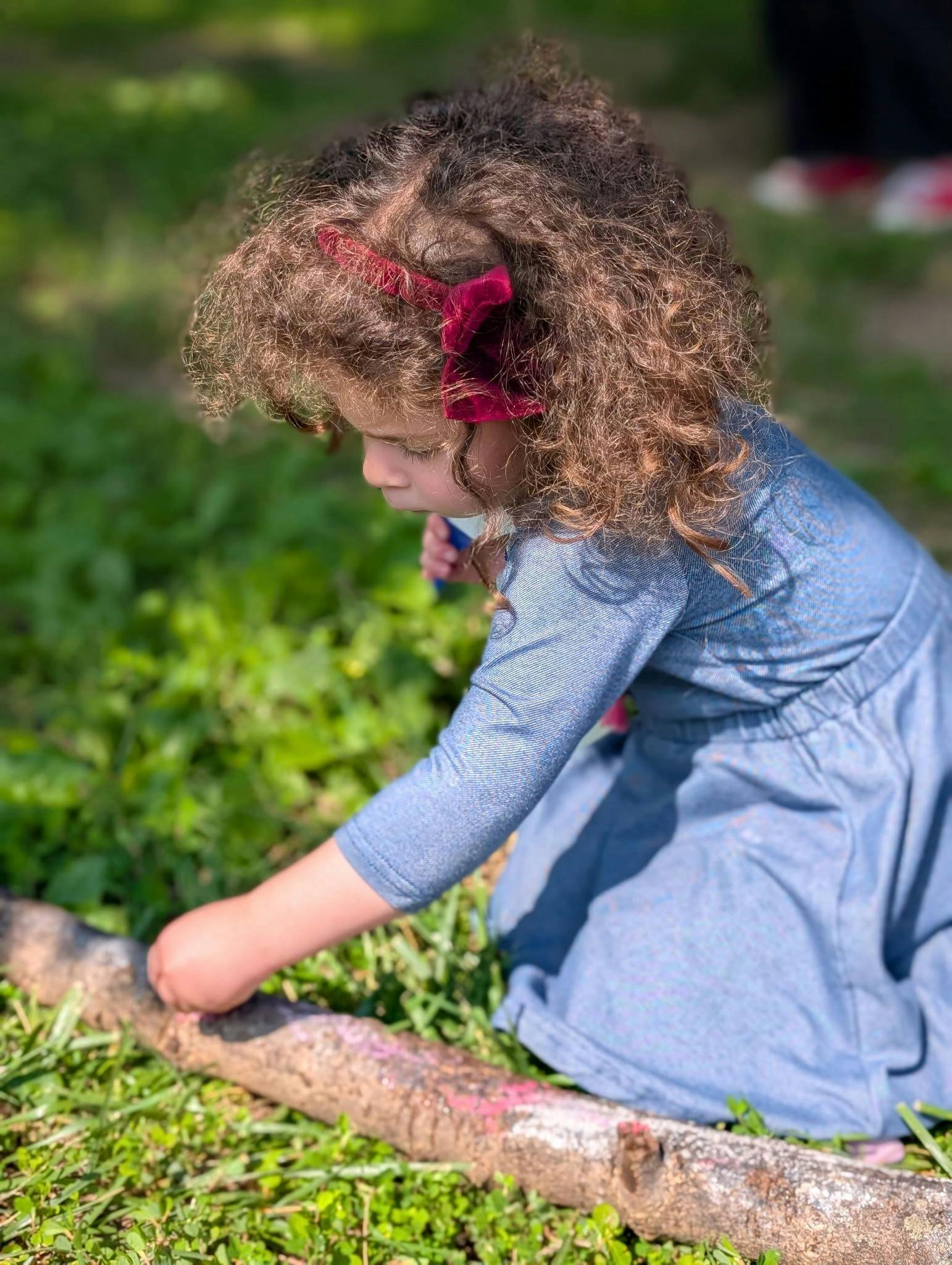 Child with curly hair and red bow touching a log on the grass
