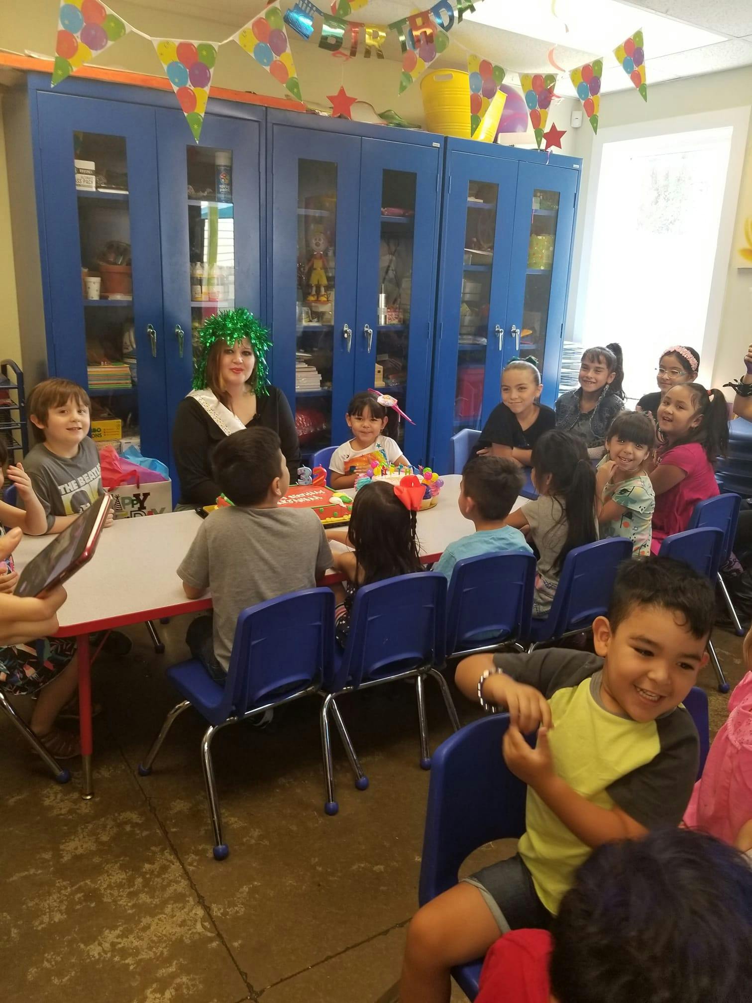 Person in green wig with children and adults at tables in a classroom