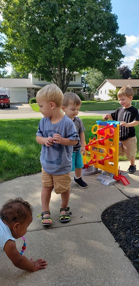 Four children playing outdoors, one crawling and three standing near a toy