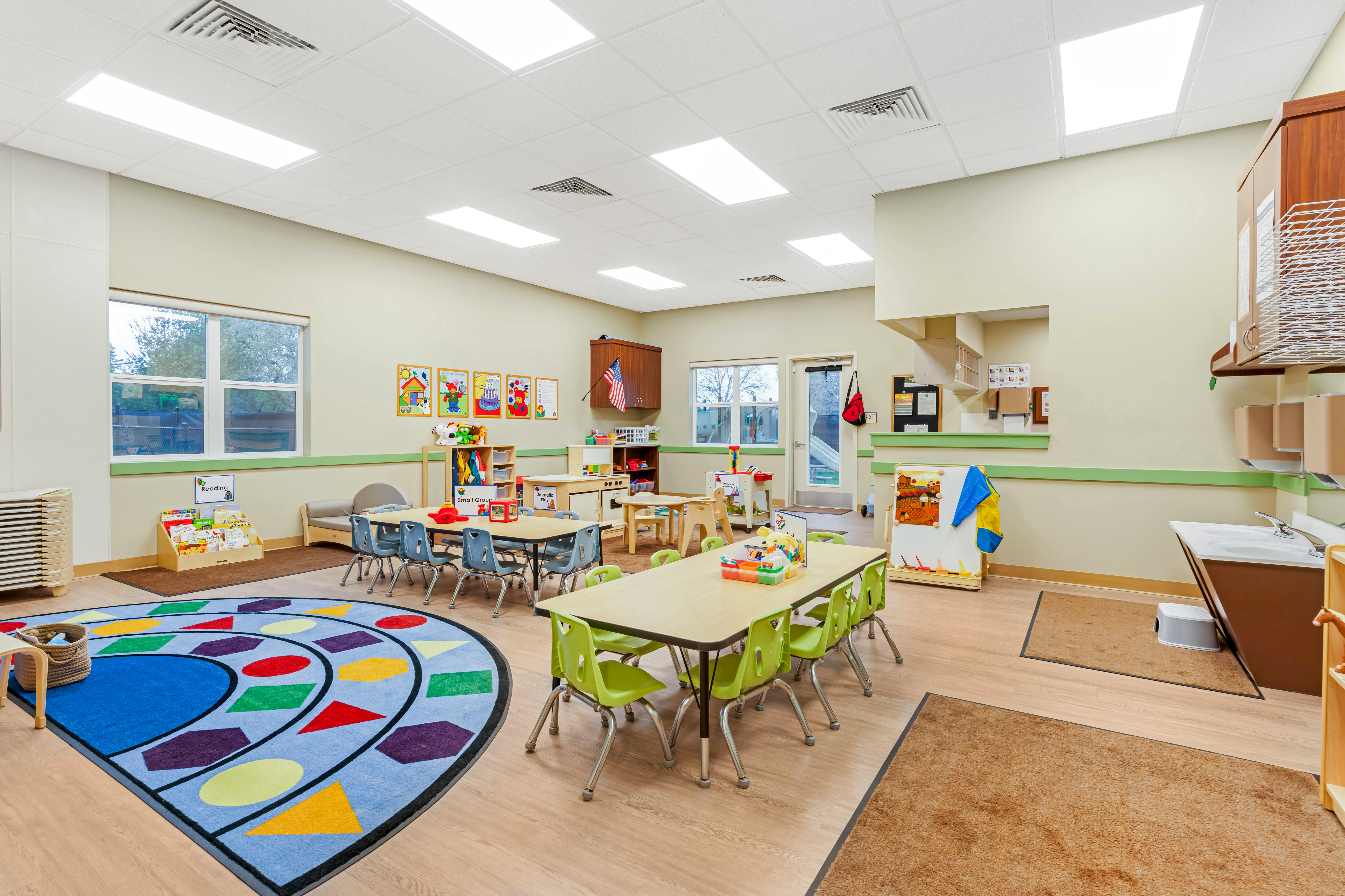An empty early childhood classroom with multiple tables and chairs, a reading corner, and a play rug