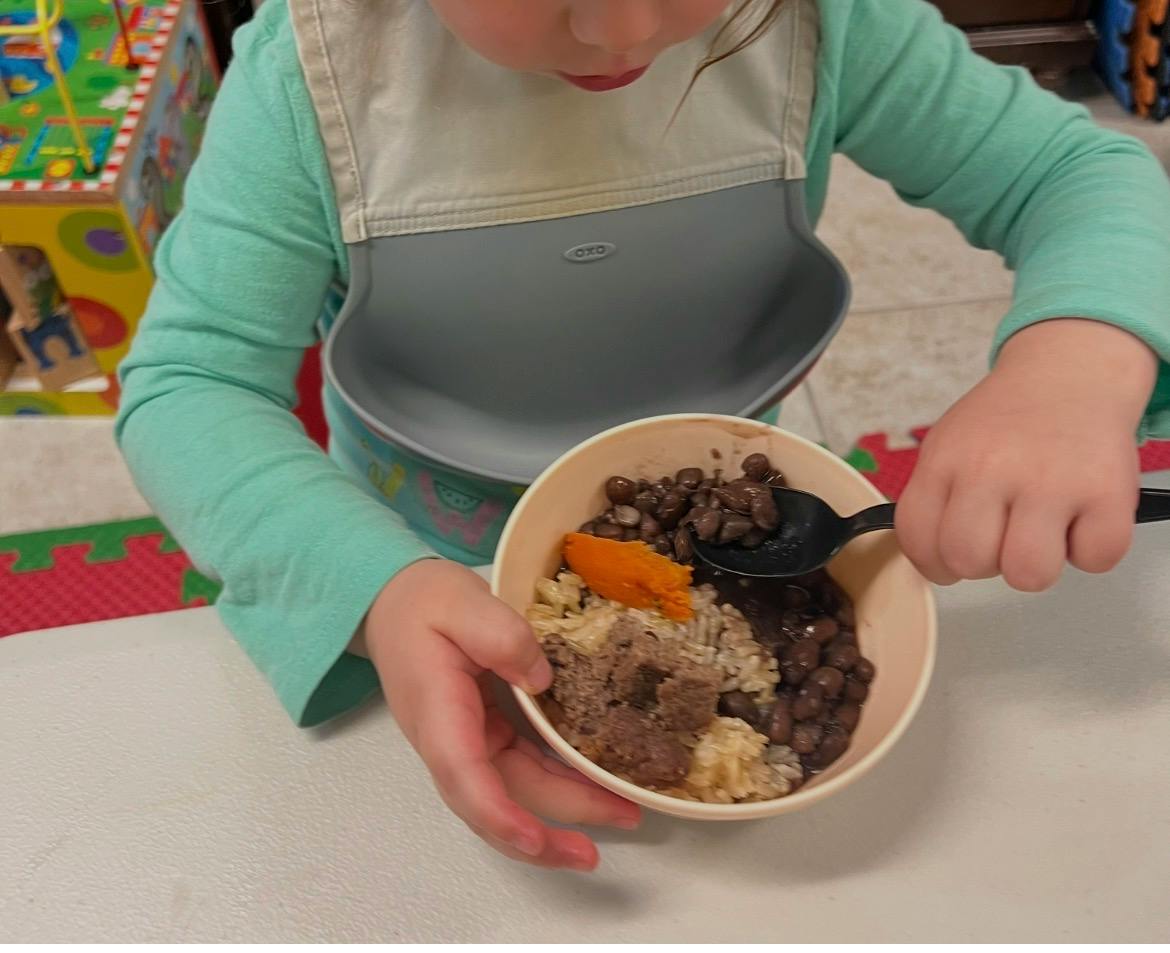 Child wearing a bib holding a bowl of food and eating with a spoon