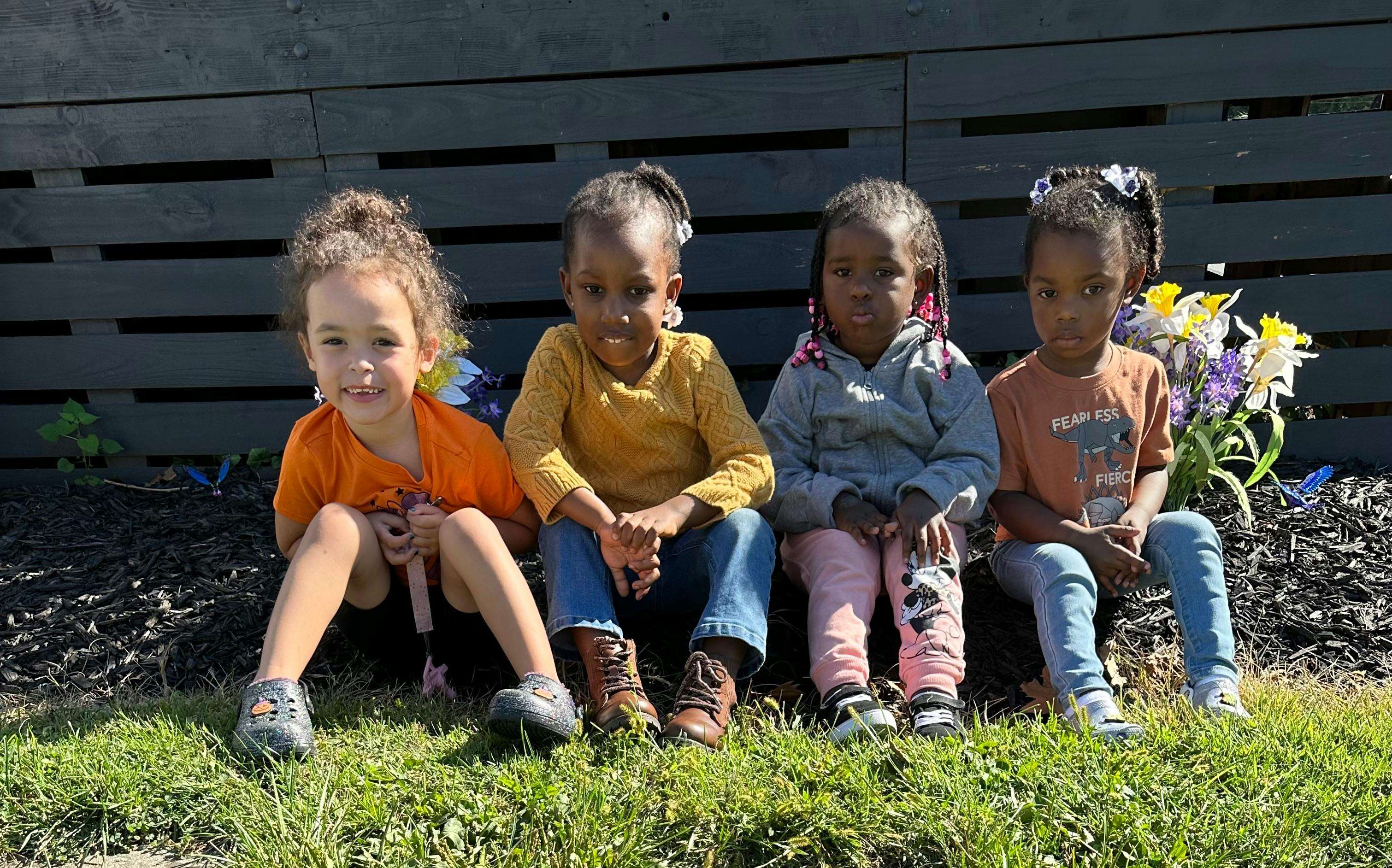 Four children sitting on grass in front of a dark fence