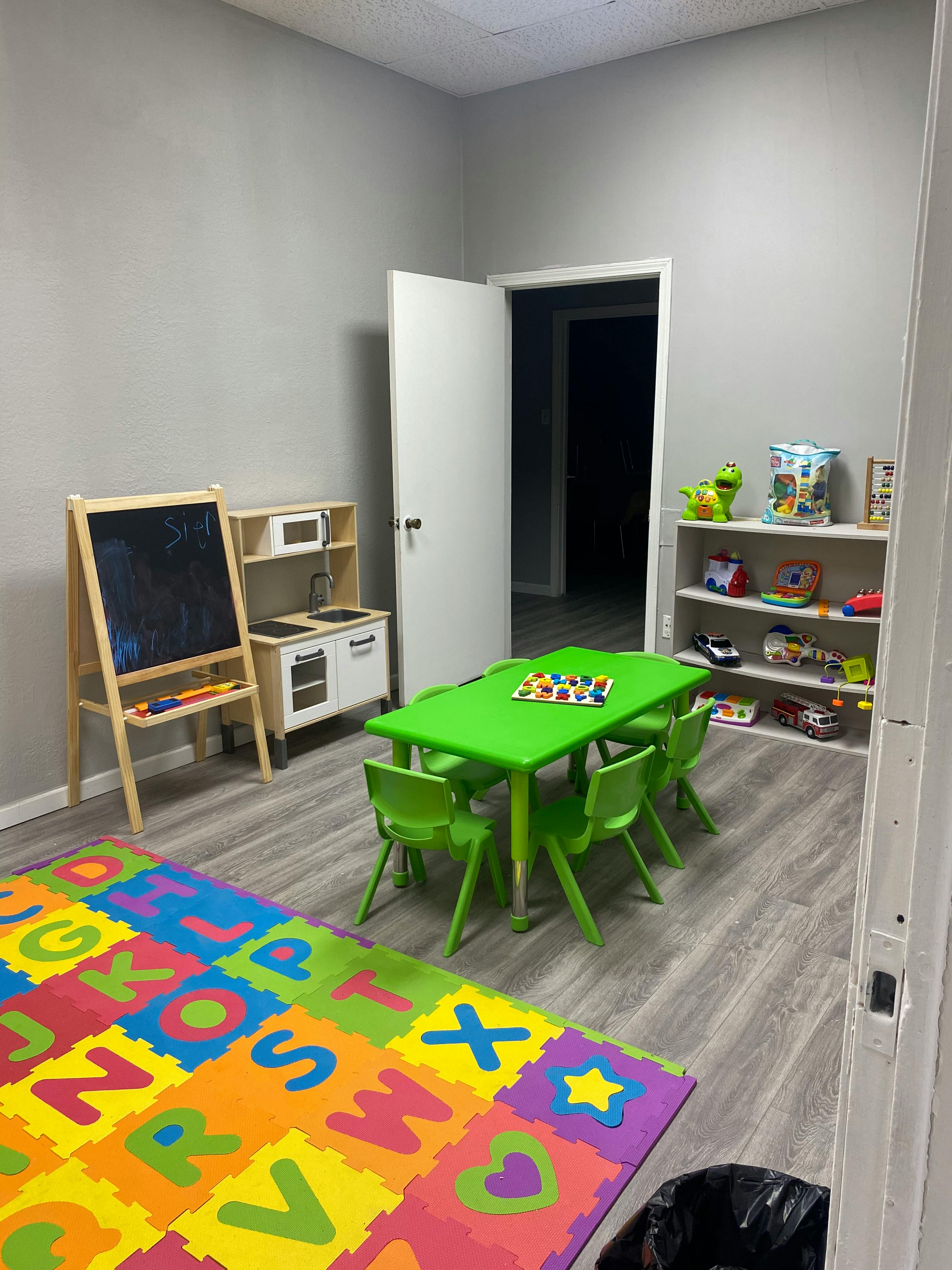 Children's play area with a green table and chairs, alphabet mat, and toy kitchen