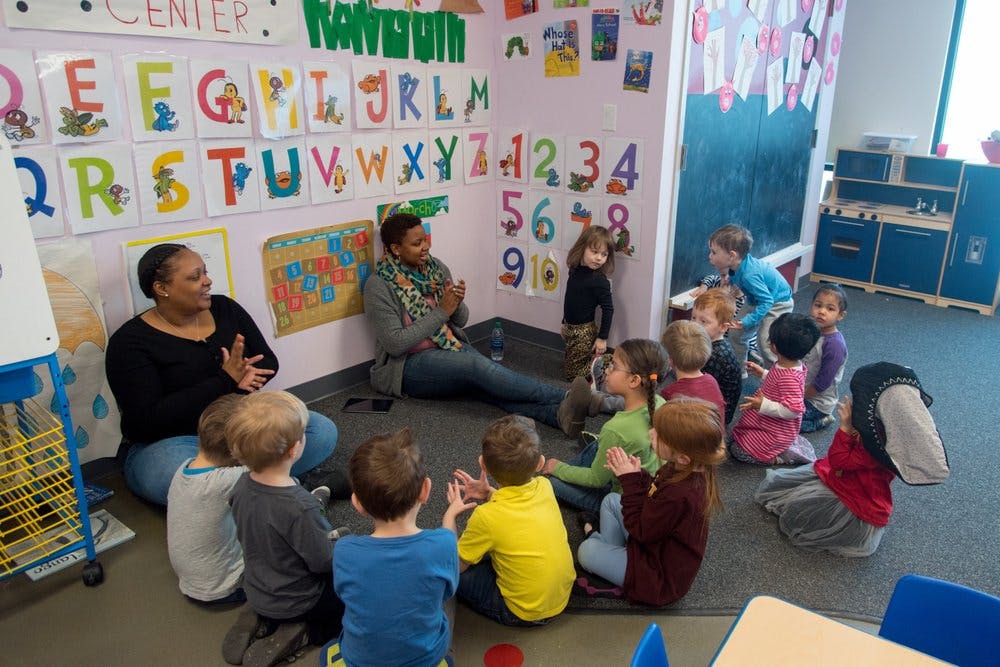 Two adults and multiple children sitting on the floor in a classroom