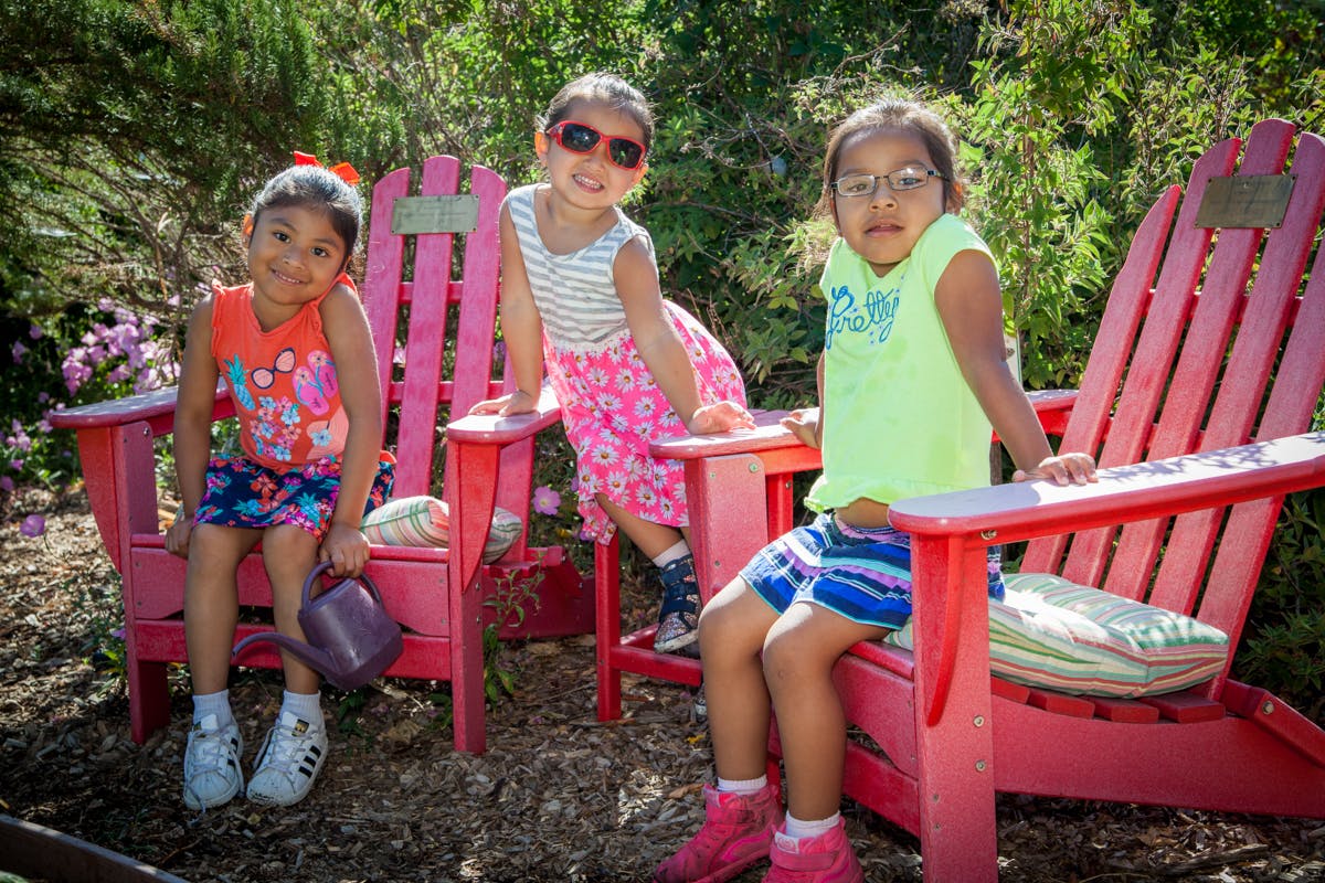 Three children sitting and standing around pink chairs outdoors