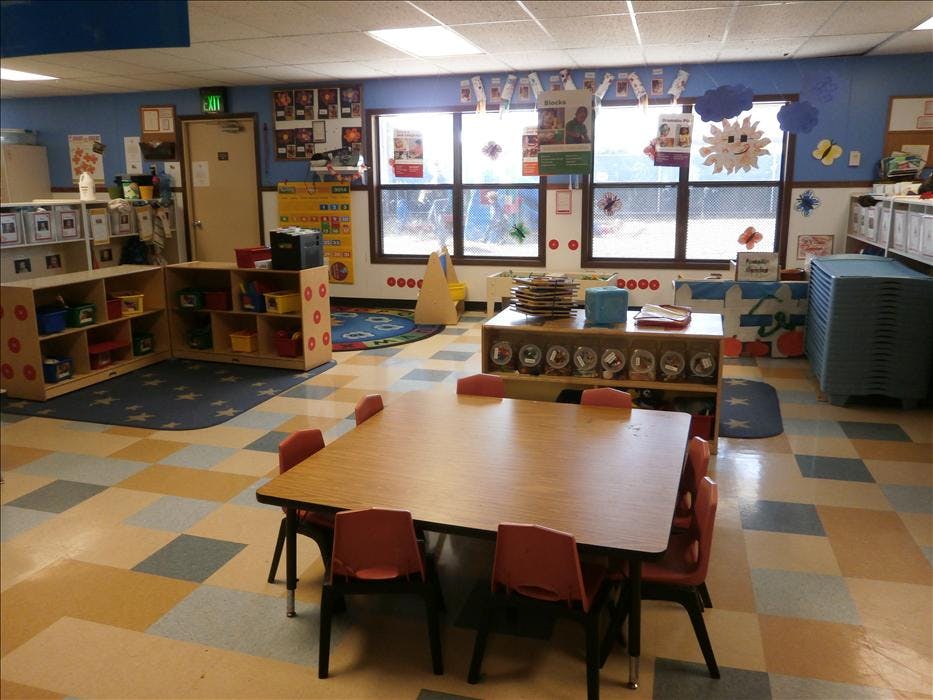 Empty elementary school classroom with tables, chairs, and shelves of supplies