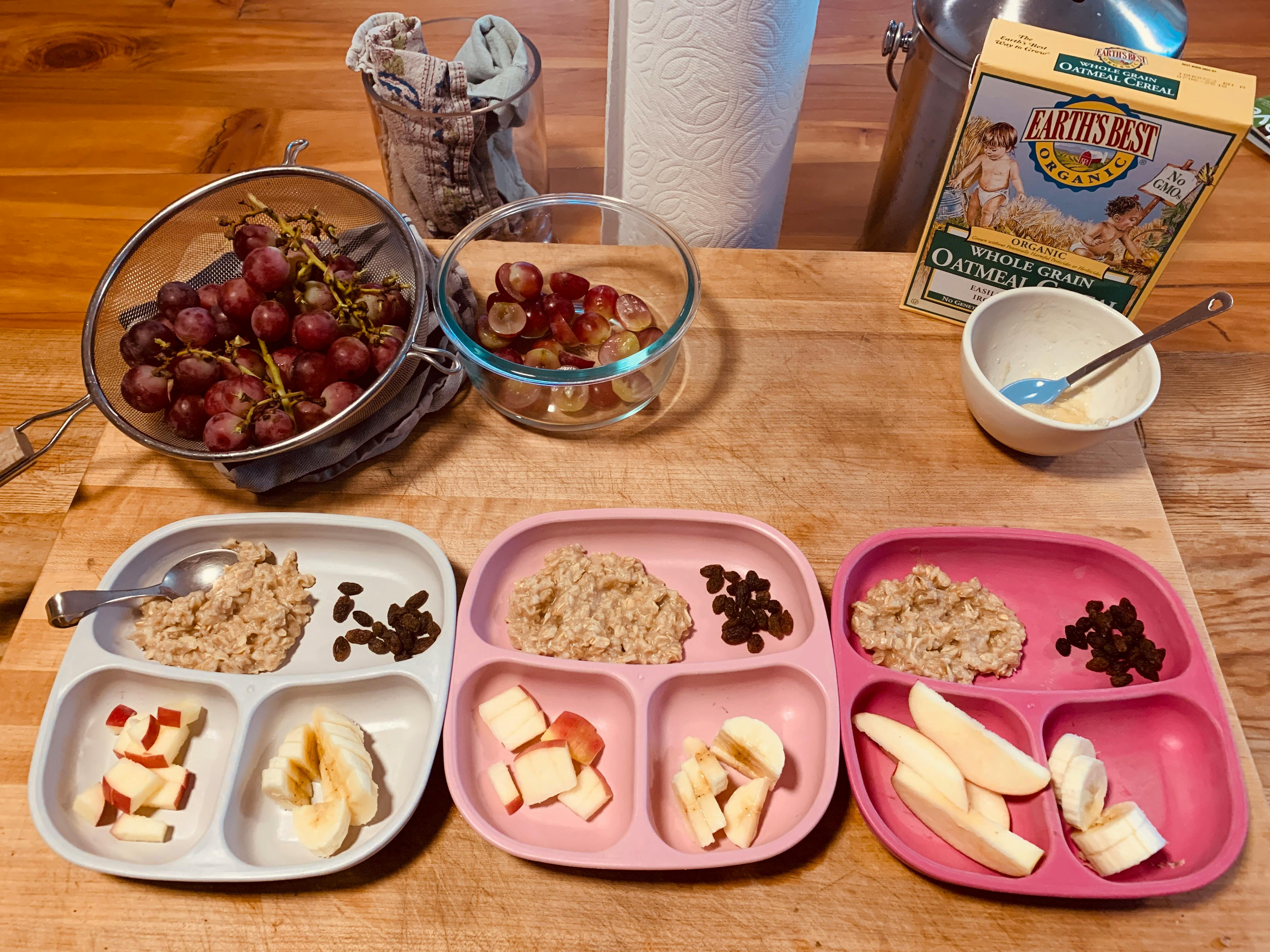Three partitioned plates with oatmeal, raisins, apples, and bananas on a wooden table
