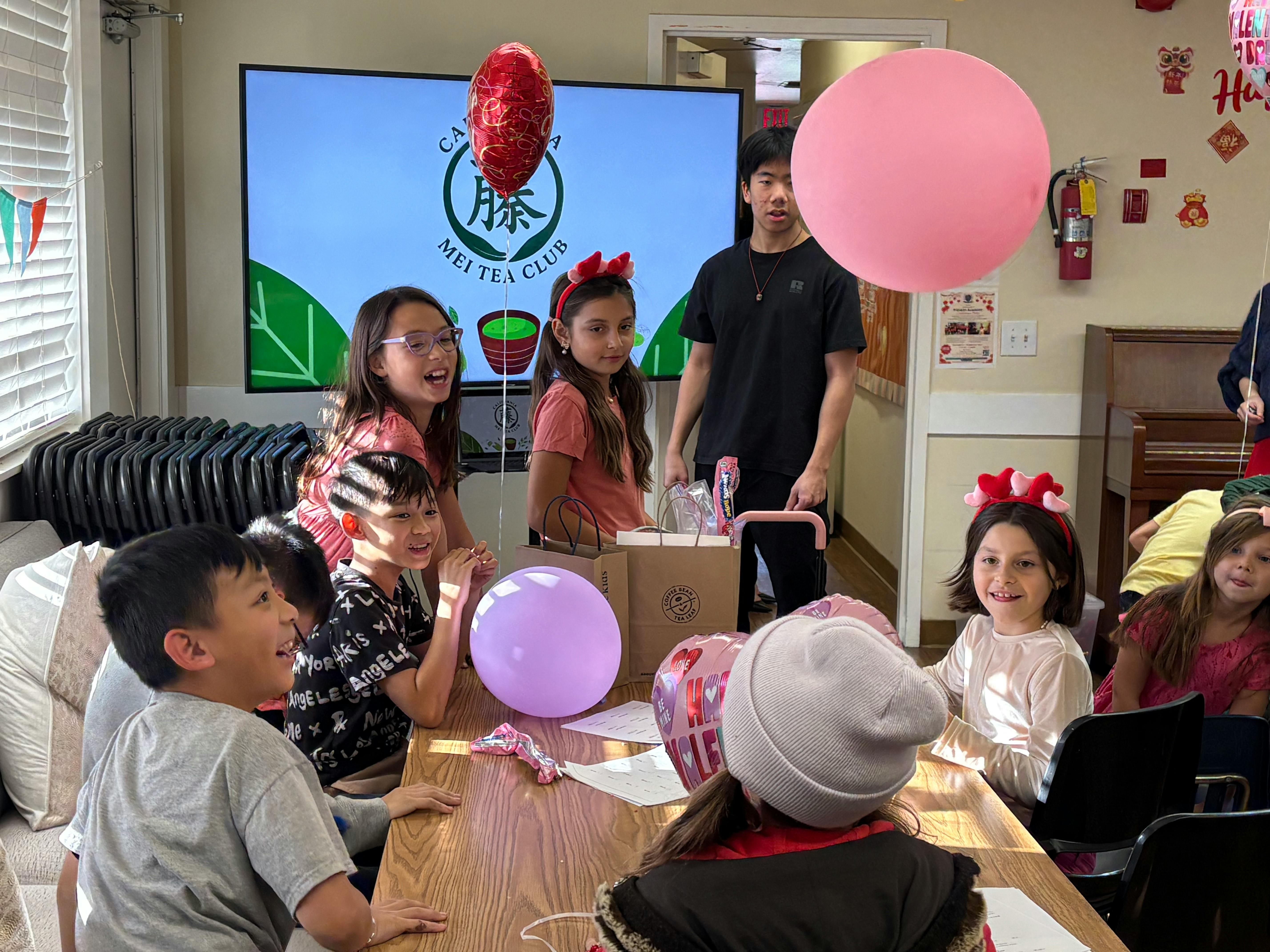 Children and a teen at a table with balloons and bags, "Mei Tea Club" on a screen