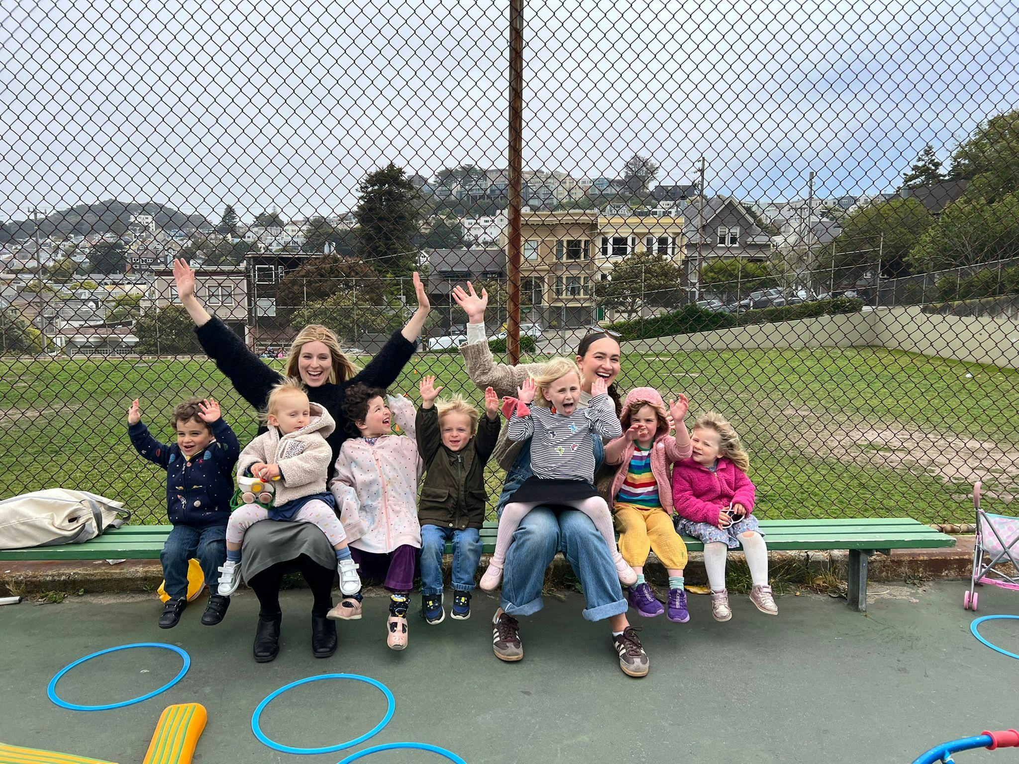 Three people holding seven children on a park bench, all waving