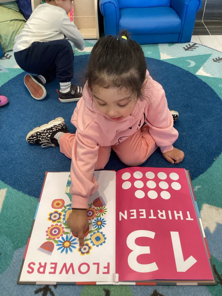 Child on floor pointing at a book with "FLOWERS" and "THIRTEEN"