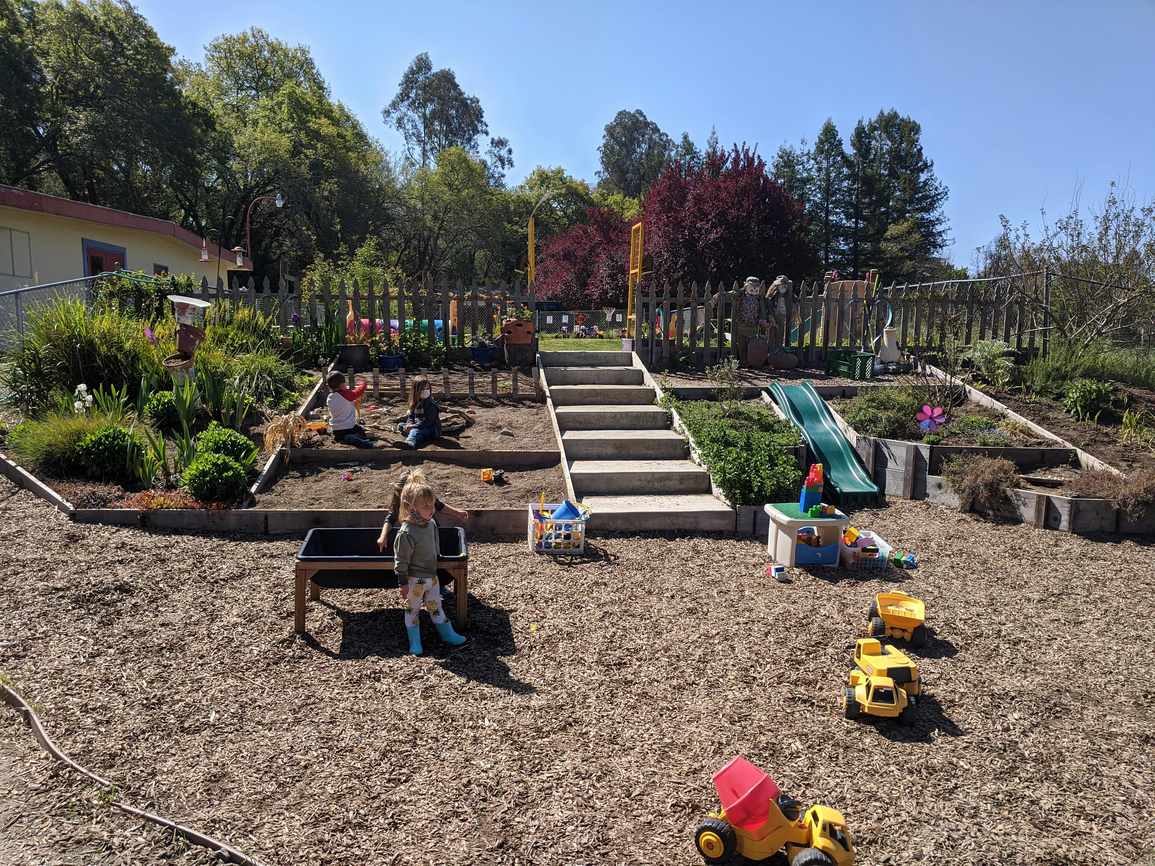 Child standing in a play area with stairs, a slide, and toy trucks