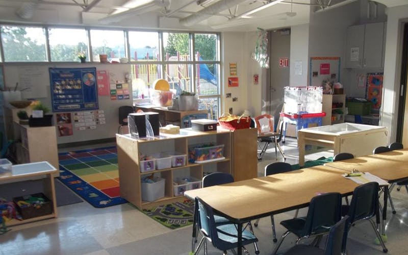 Classroom with tables, shelves, colorful rug, and a view of an outdoor playground
