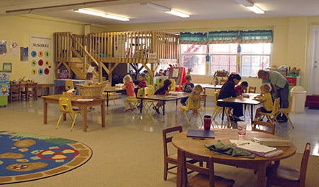 Children and adults in a classroom with tables and a wooden play structure