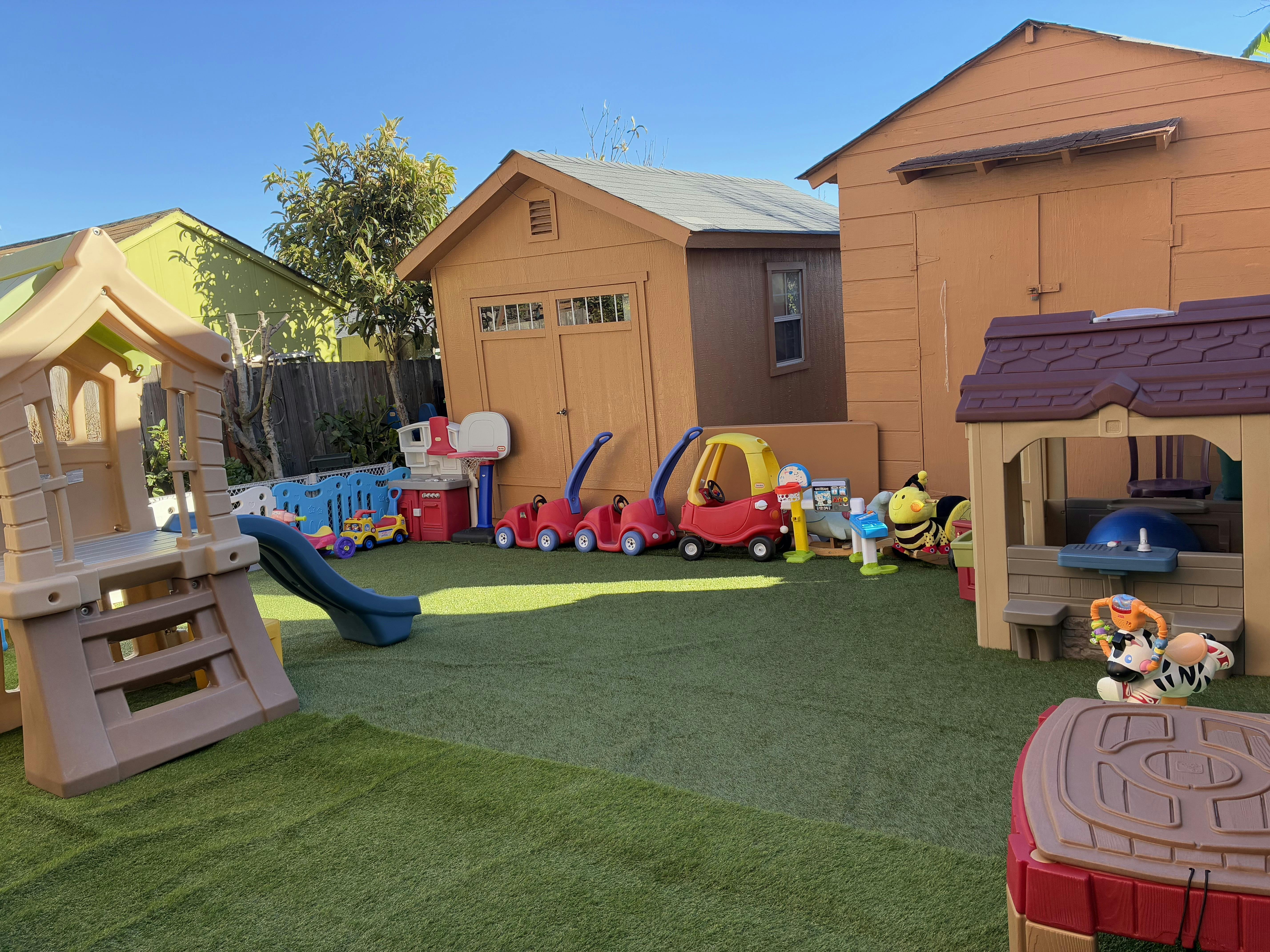 Plastic play structure with slide, toy cars, and sheds in a yard with artificial grass