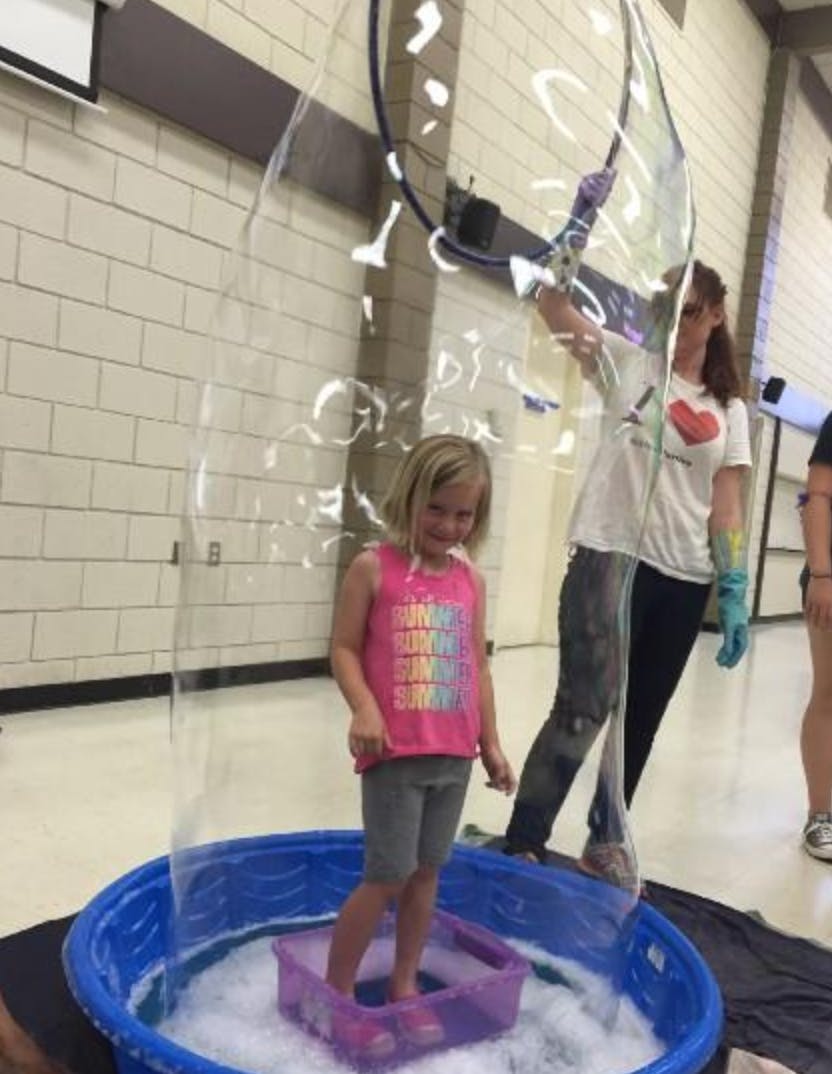 Child standing in a bubble solution while a person creates a large bubble around them