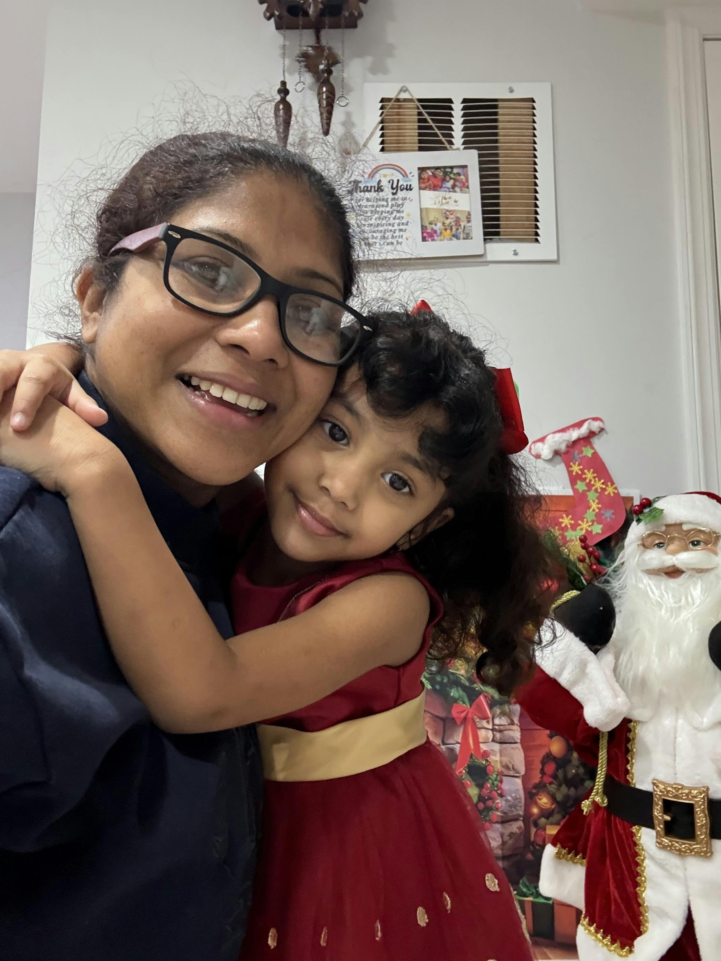 Person in glasses embracing a child in a red dress, Santa figure on the right