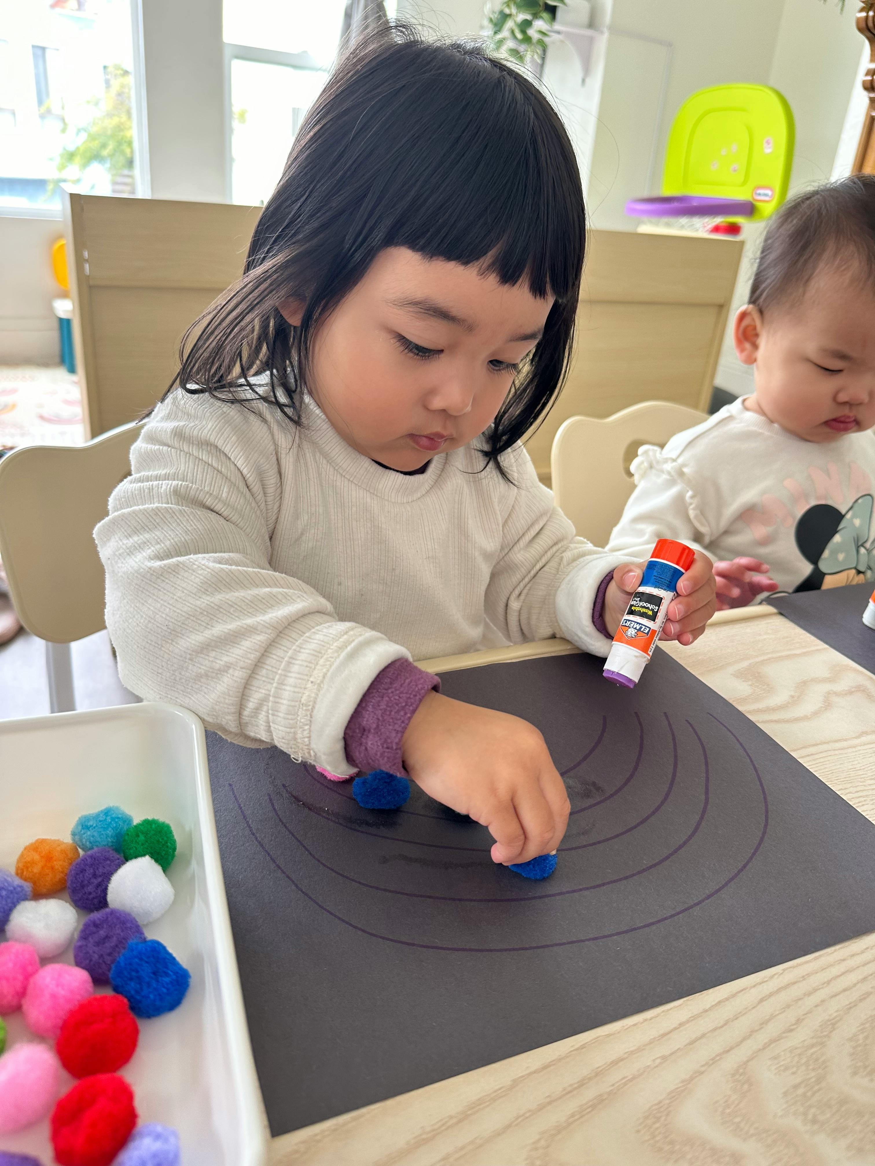 Child gluing pom-poms onto a black paper with another child in the background