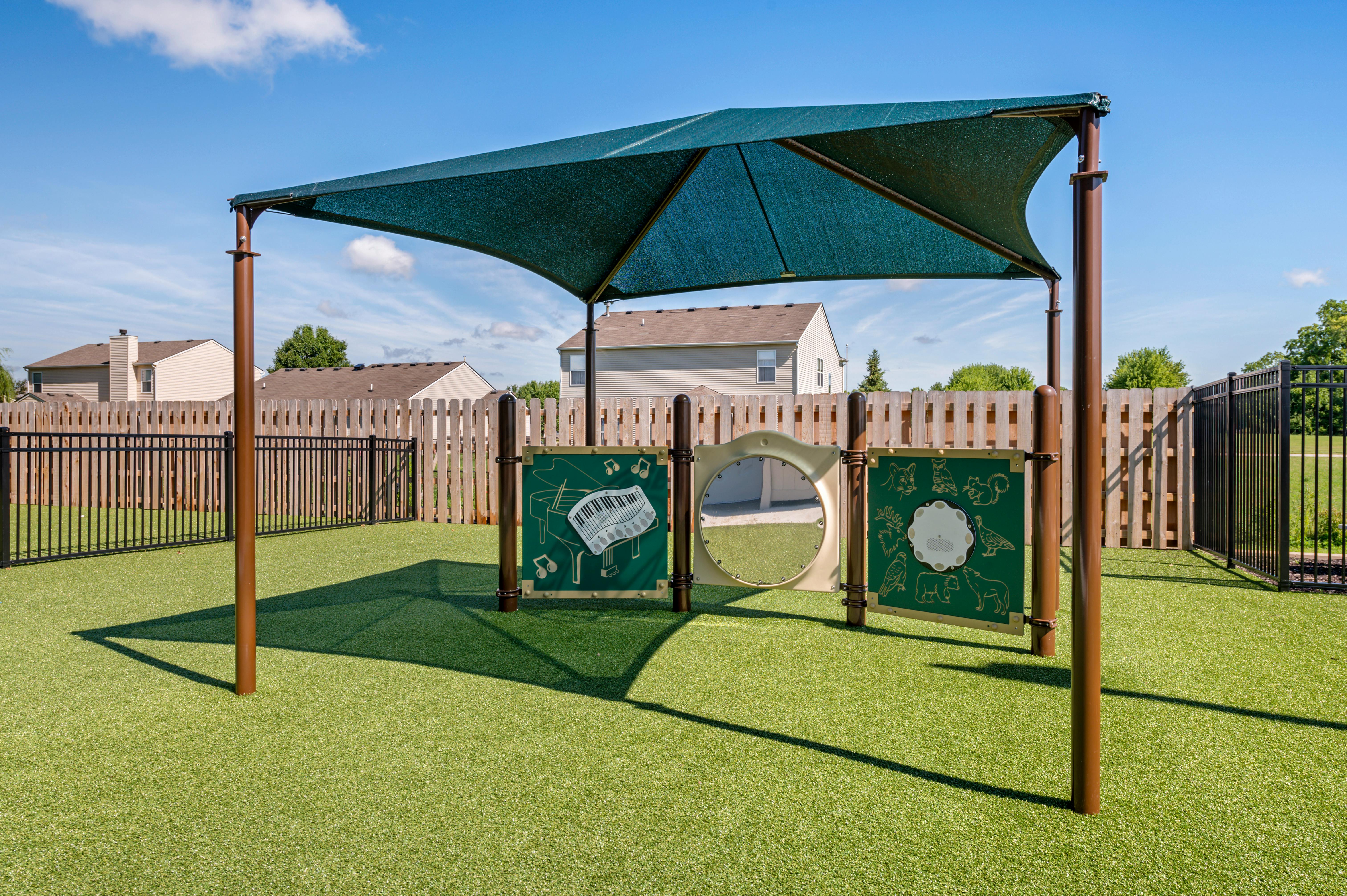 A playground with a green canopy and interactive panels on artificial turf