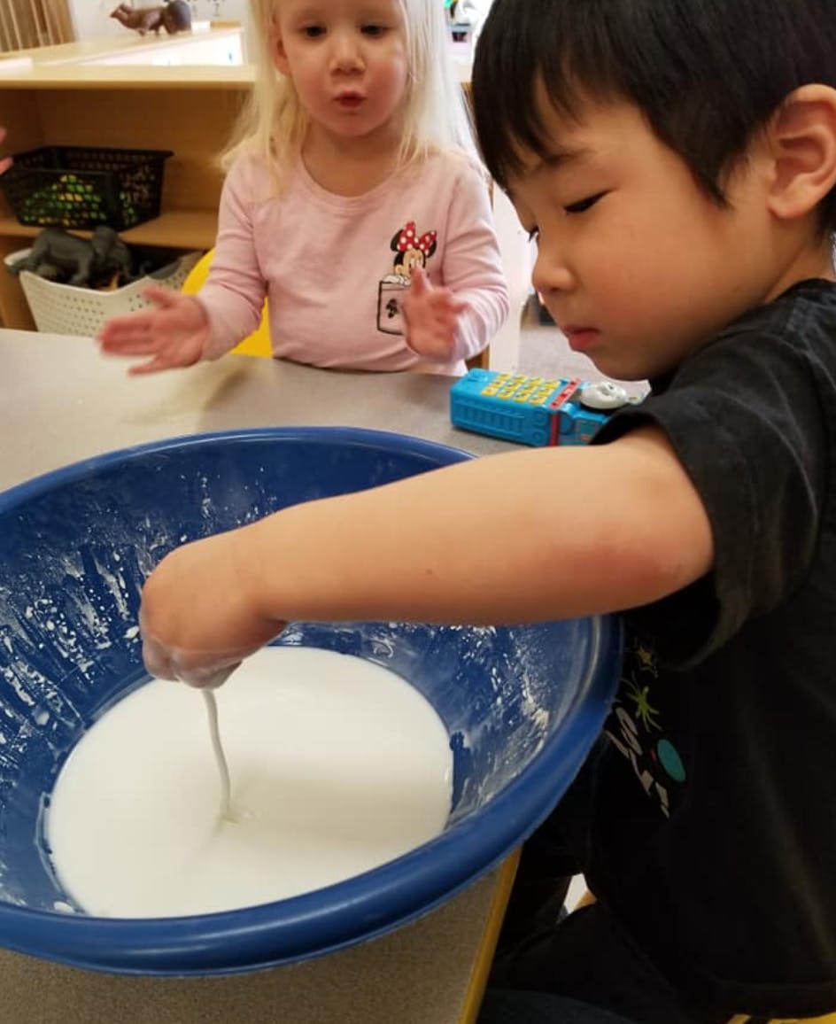 Child mixing white liquid in a blue bowl, another child watching