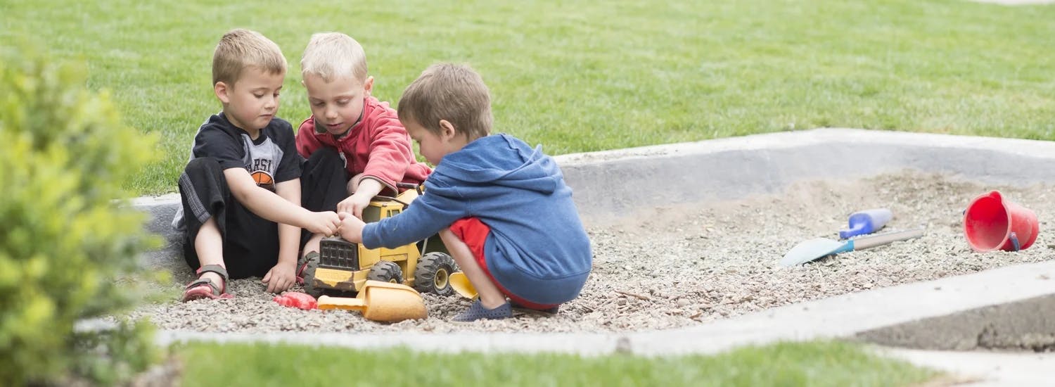 Three children playing with toy trucks in a sandpit