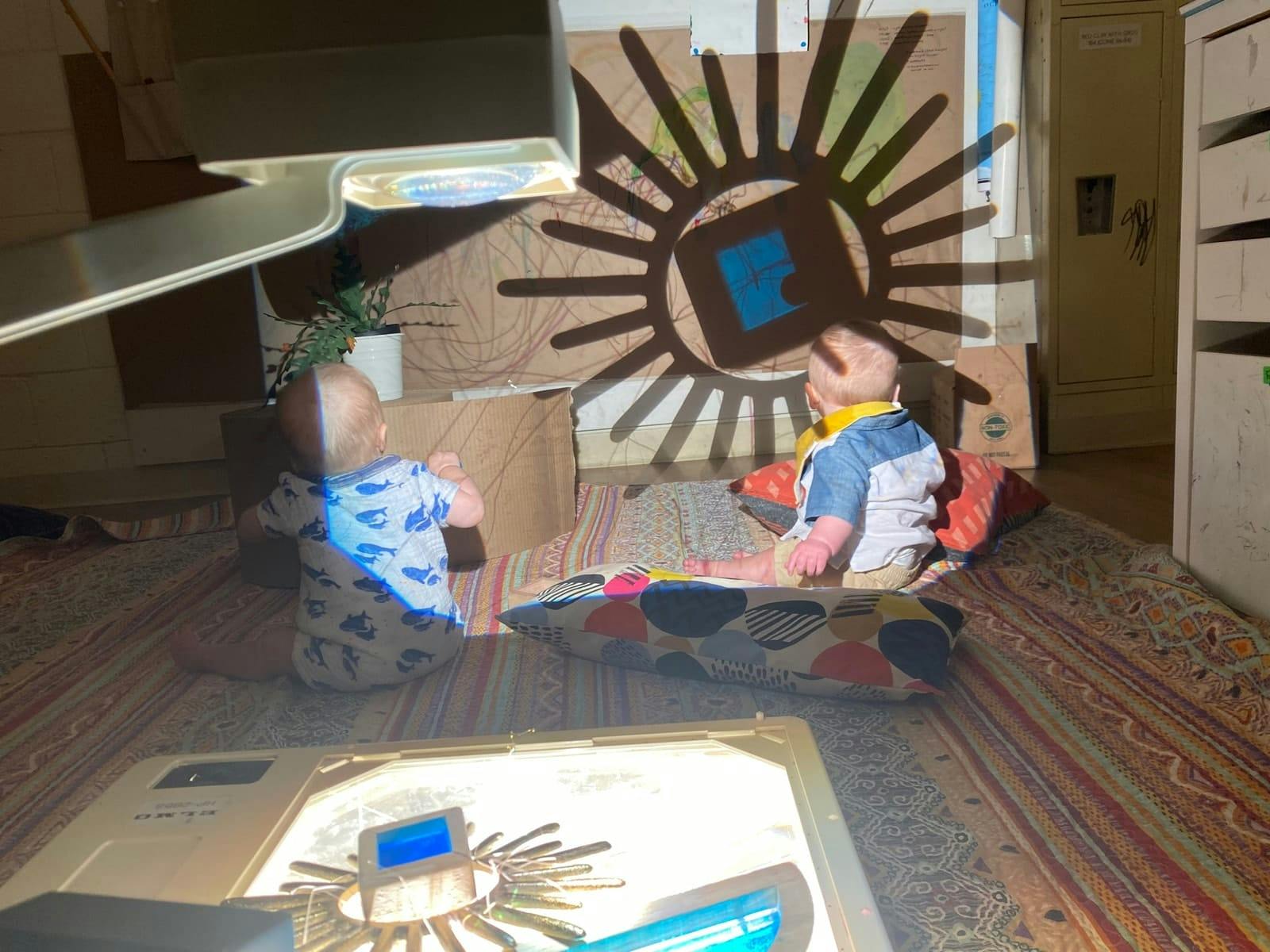 Two children sitting on a rug watching a sun-shaped shadow projection