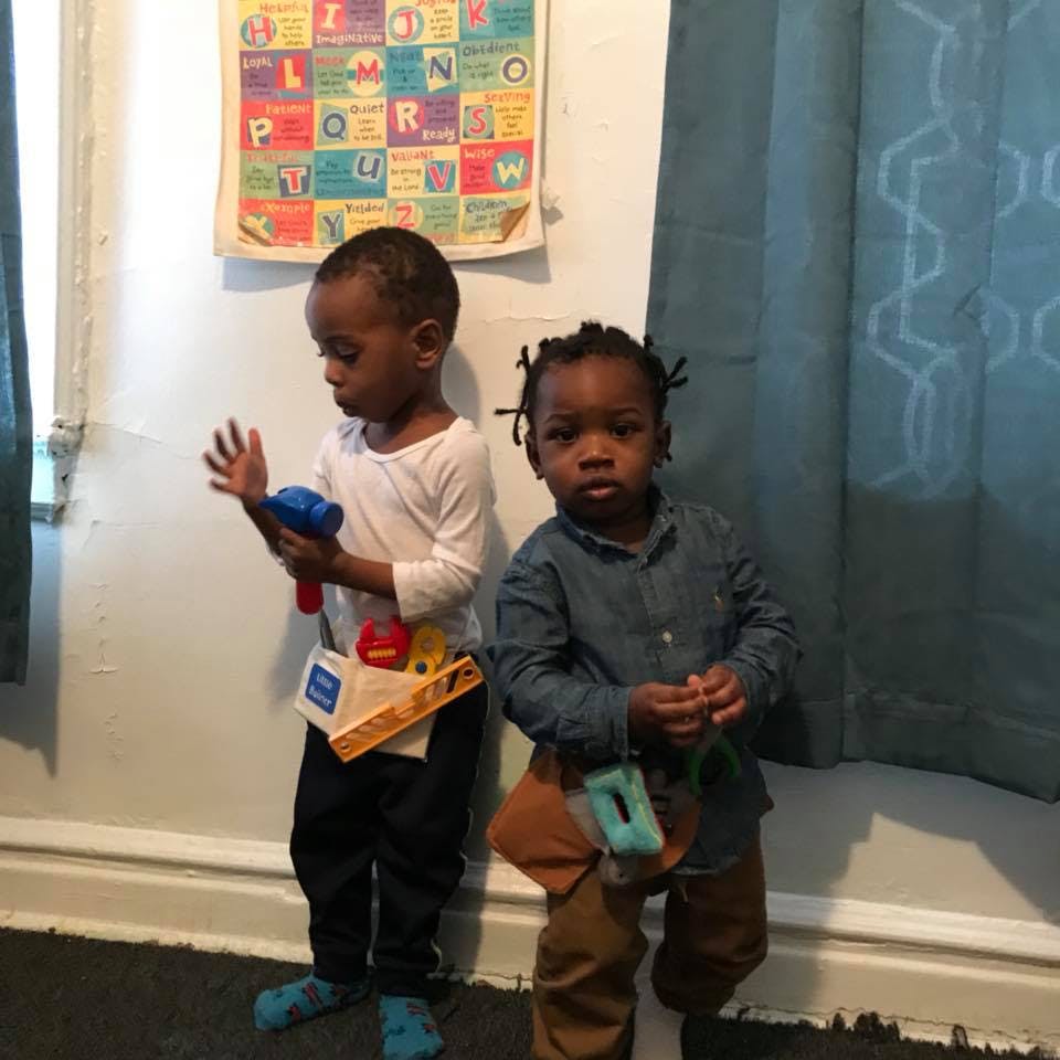Two children standing, one holding a toy hammer and the other wearing a toy tool belt